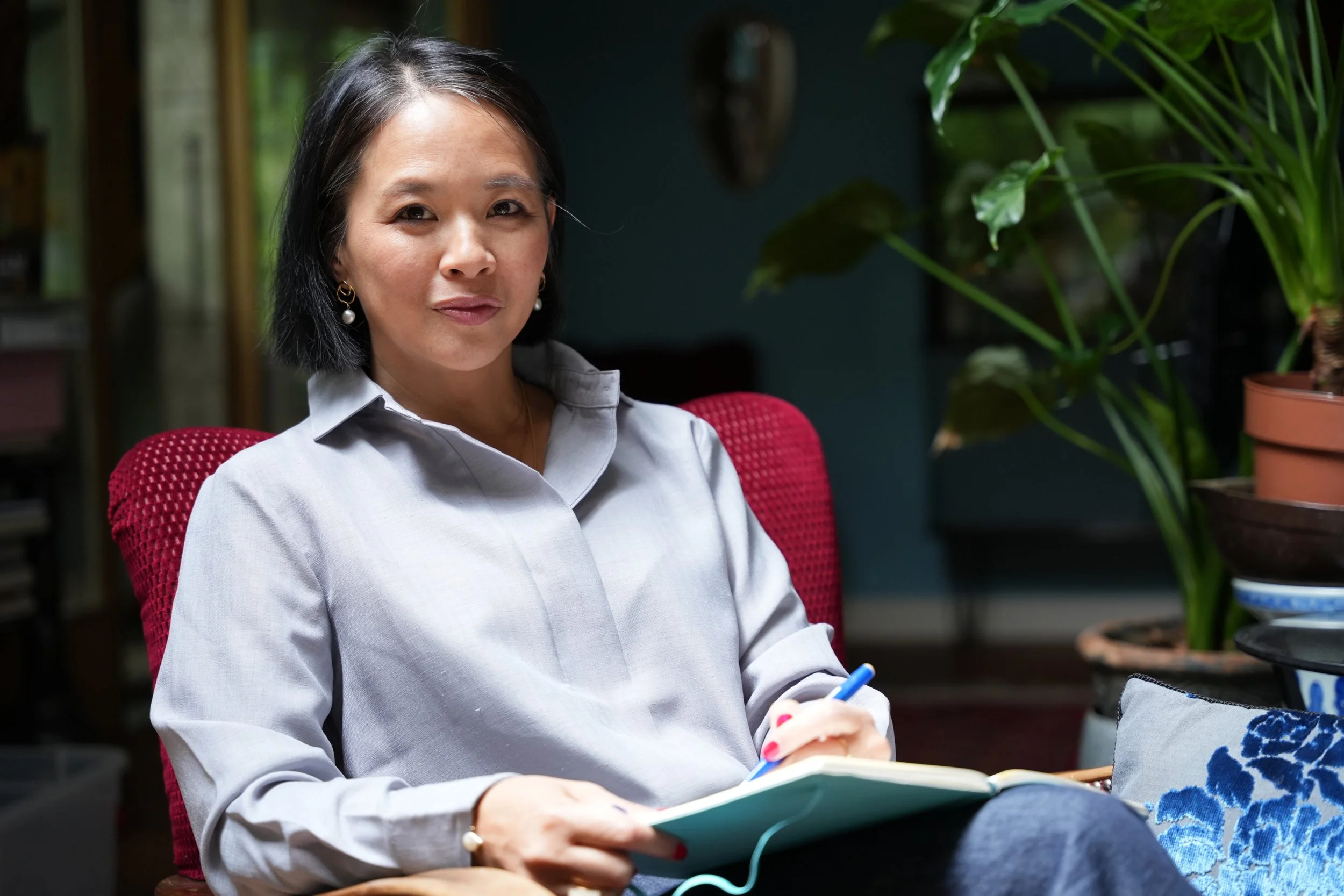 Femme assise, tenant un stylo et un carnet, dans un intérieur avec des plantes. Yen-Nhi Nguyen - coach d'exception - coach de dirigeants -cercles de dirigeants - accompagner les dirigeants - leaadership - management - formations - cerveau - intellige