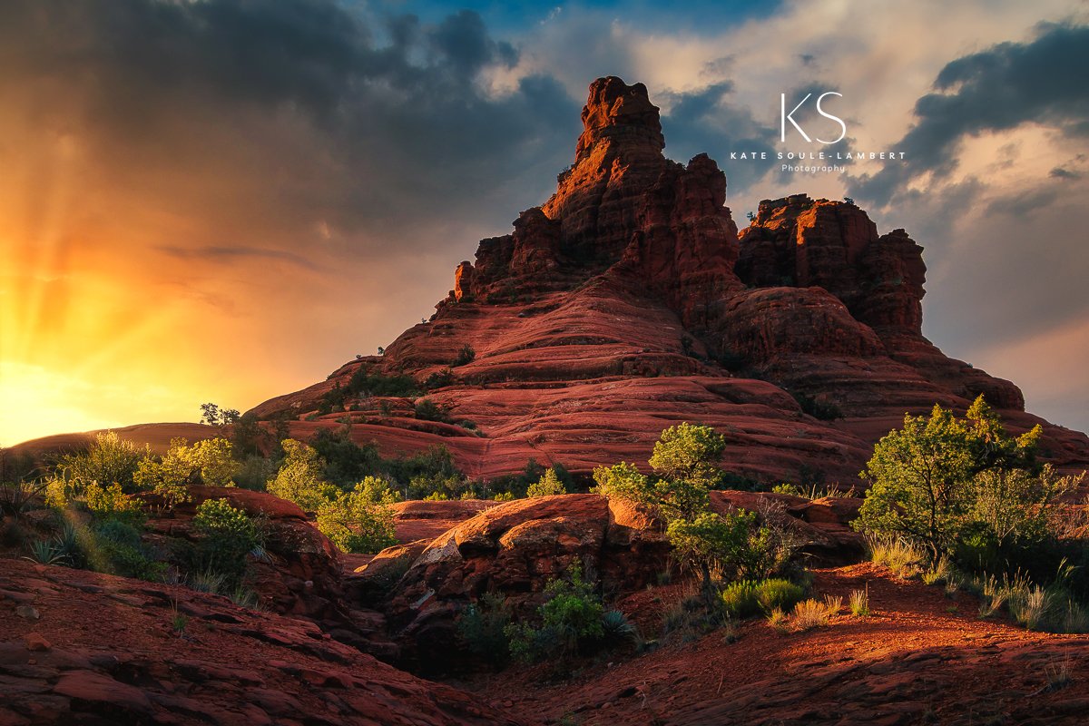 Sunset over a large red rock formation with green trees in the foreground and a sky with clouds.