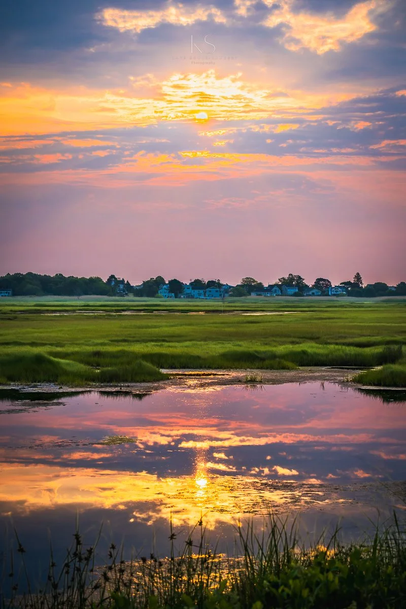 Sunset over a marsh with a reflected sky and clouds, grassy foreground, and distant houses under a colorful sky.