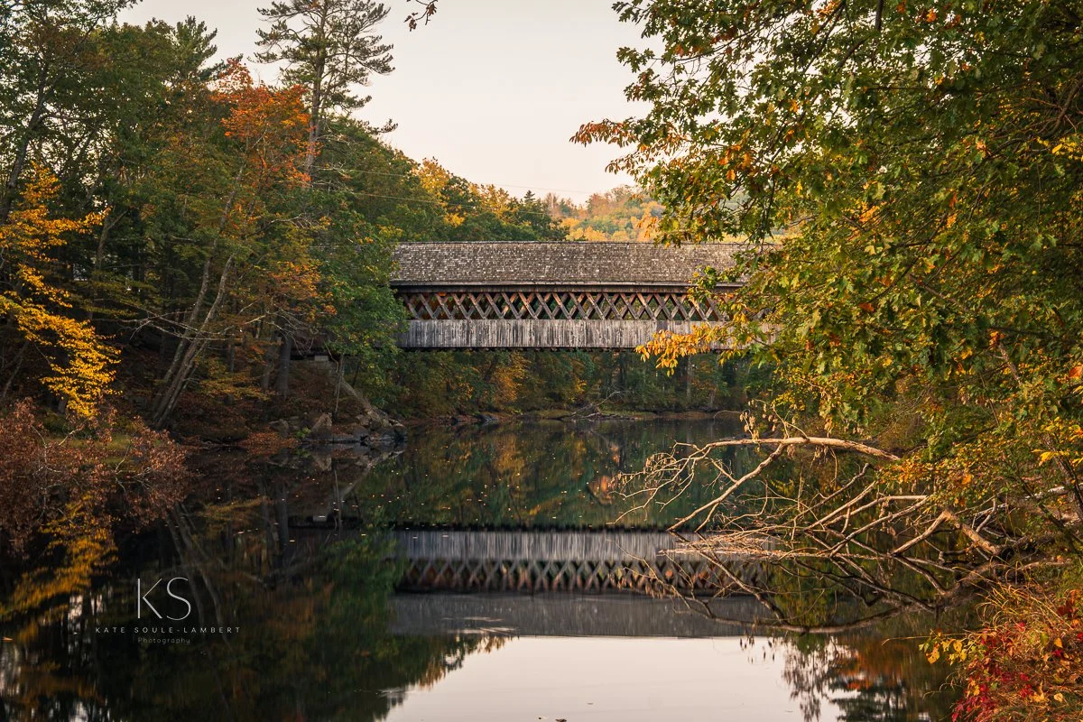 A covered wooden bridge over a calm river, surrounded by trees with autumn foliage.