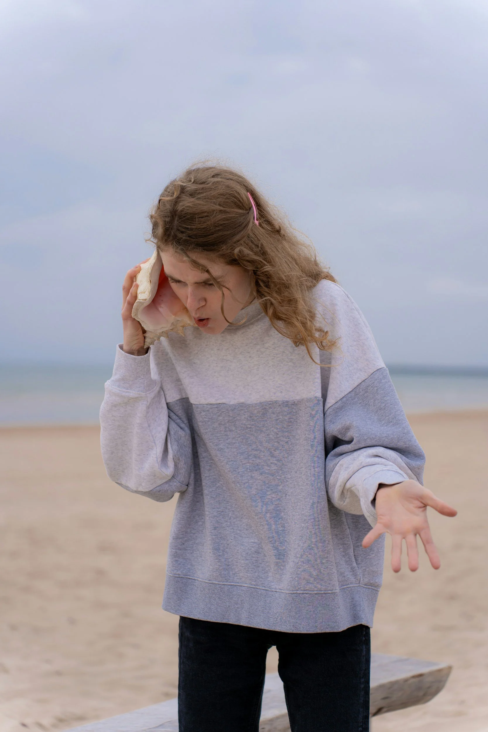Young girl at the beach holding a large seashell to her ear and appearing to listen, with an overcast sky and ocean in the background.