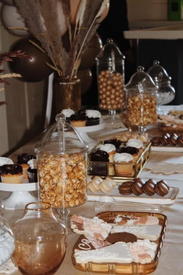 Table set with various desserts and candies, including cupcakes with frosting, caramel popcorn in glass jars, assorted cookies, and chocolates.