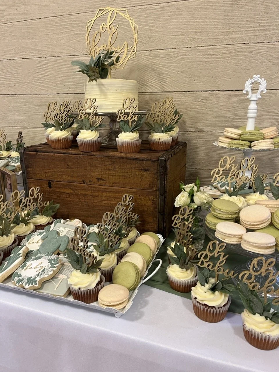 Dessert table with cupcakes, macarons, and cookies decorated with "Oh Baby" signs and greenery at a celebration.
