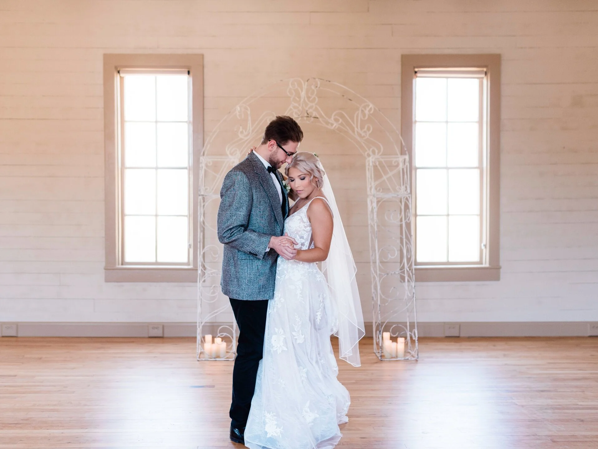 A bride and groom holding hands and leaning close during their wedding ceremony in a bright room with wood floors, large windows, and lit candles in the background.
