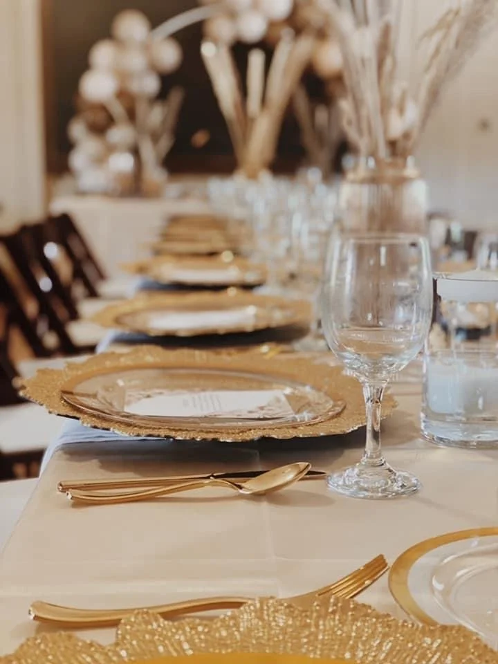 Elegant dining table with gold and cream-colored plates, gold cutlery, a clear wine glass, and decorative centerpieces with dried flowers.