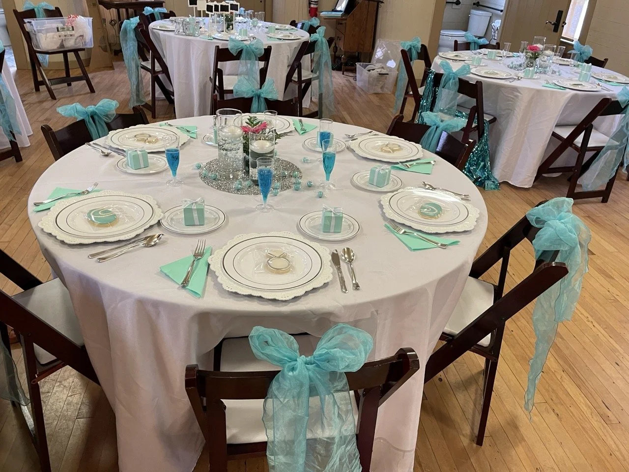 Round banquet table decorated with white tablecloth, teal napkins, and teal tulle bows on dark wooden chairs. Place settings include white plates, silverware, and small teal gift boxes. Centerpiece features candles and teal and silver accents.