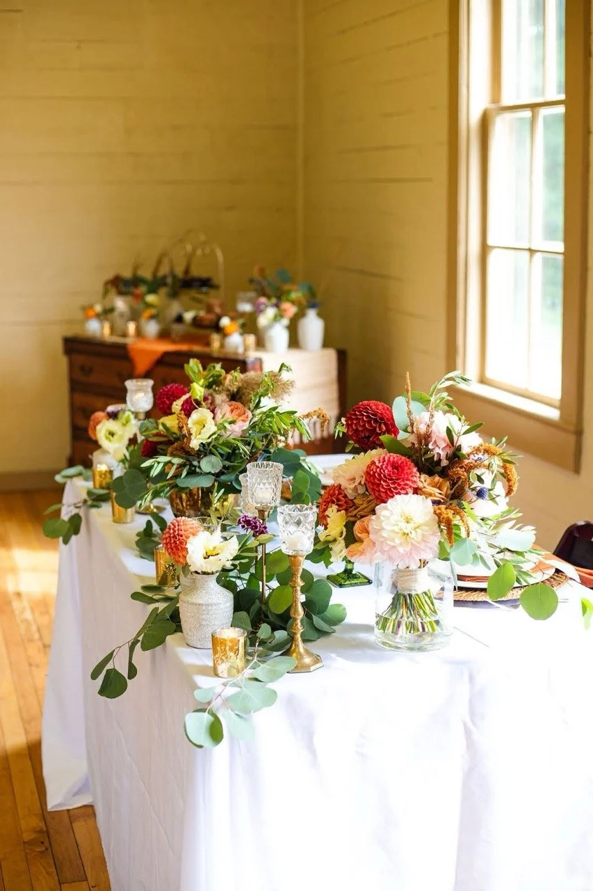 A table decorated with floral arrangements, greenery, and candles in a bright room with wooden floors and large windows.