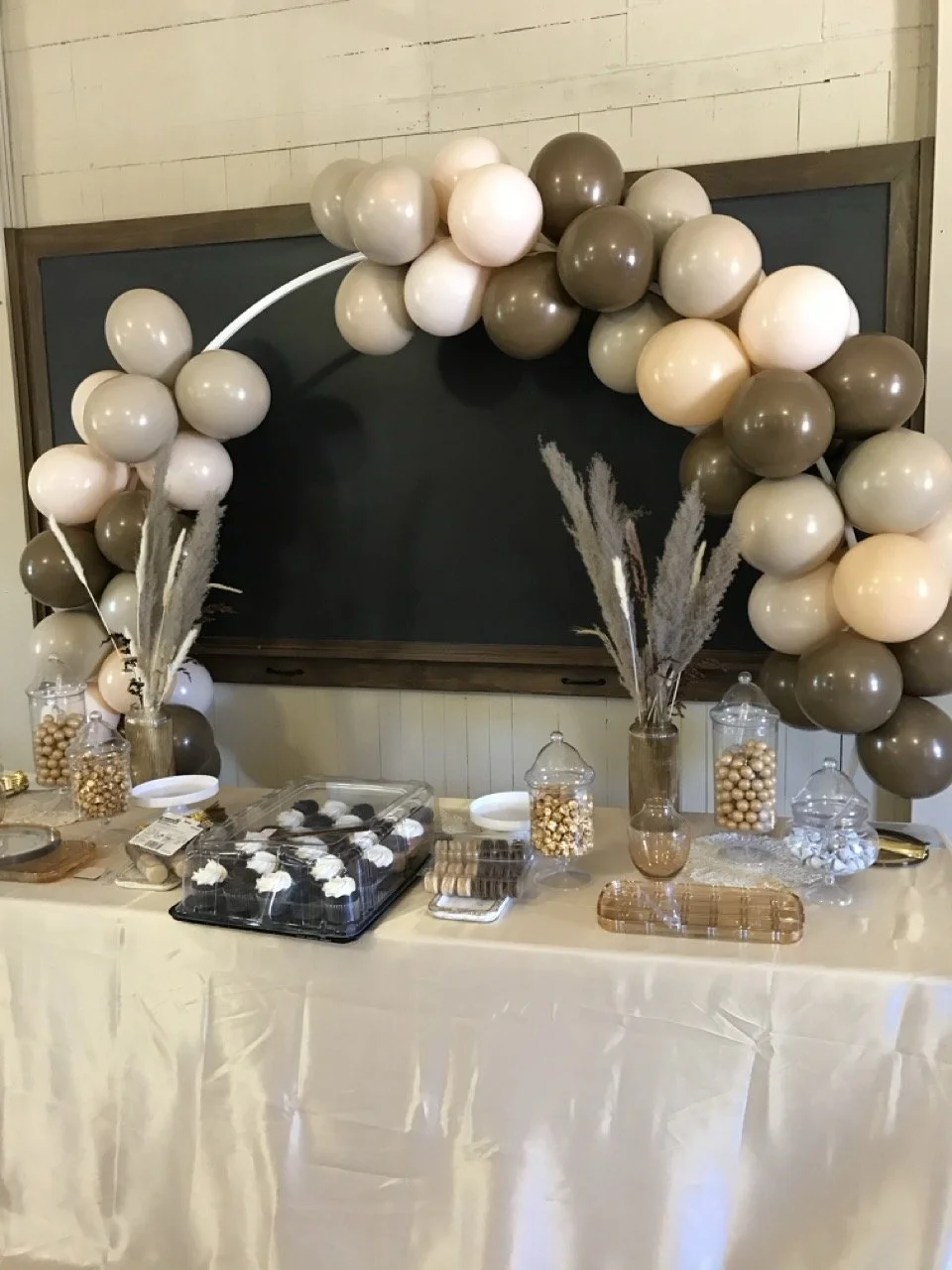 Dessert table decorated with beige, white, and brown balloons, with jars of candies and cookies, and dried floral arrangements in vases.