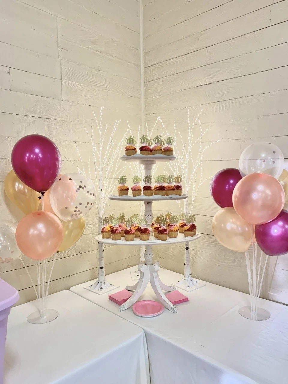 Party display with cupcakes on a white tiered stand, surrounded by pink, gold, and white balloons, and white decorative branches in the background.