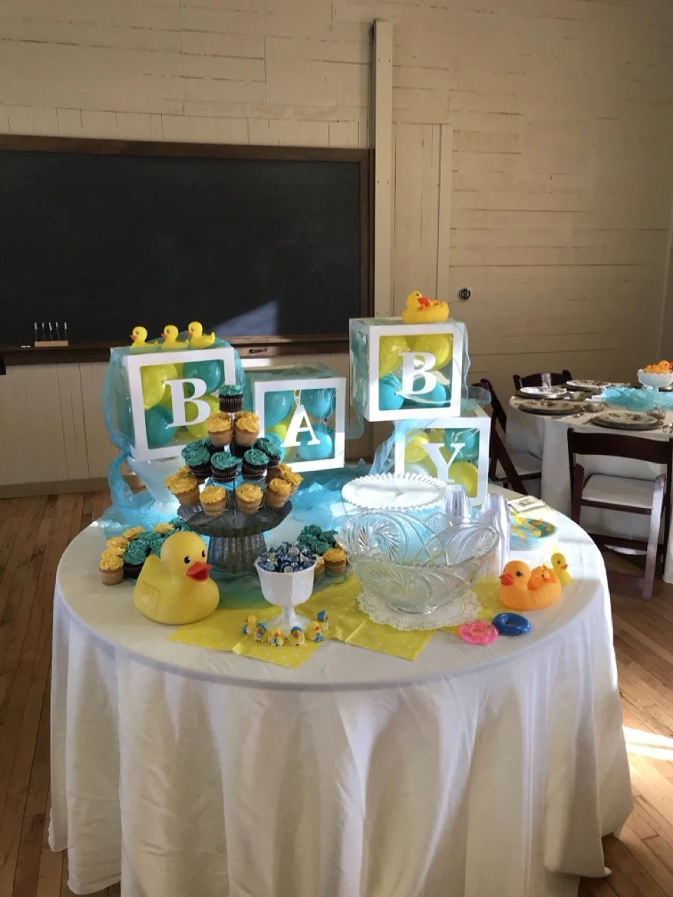 Baby shower dessert table with yellow and blue decorations, rubber duck toys, cupcakes, candy, and lighted blocks displaying the letters B, A, B, and Y.