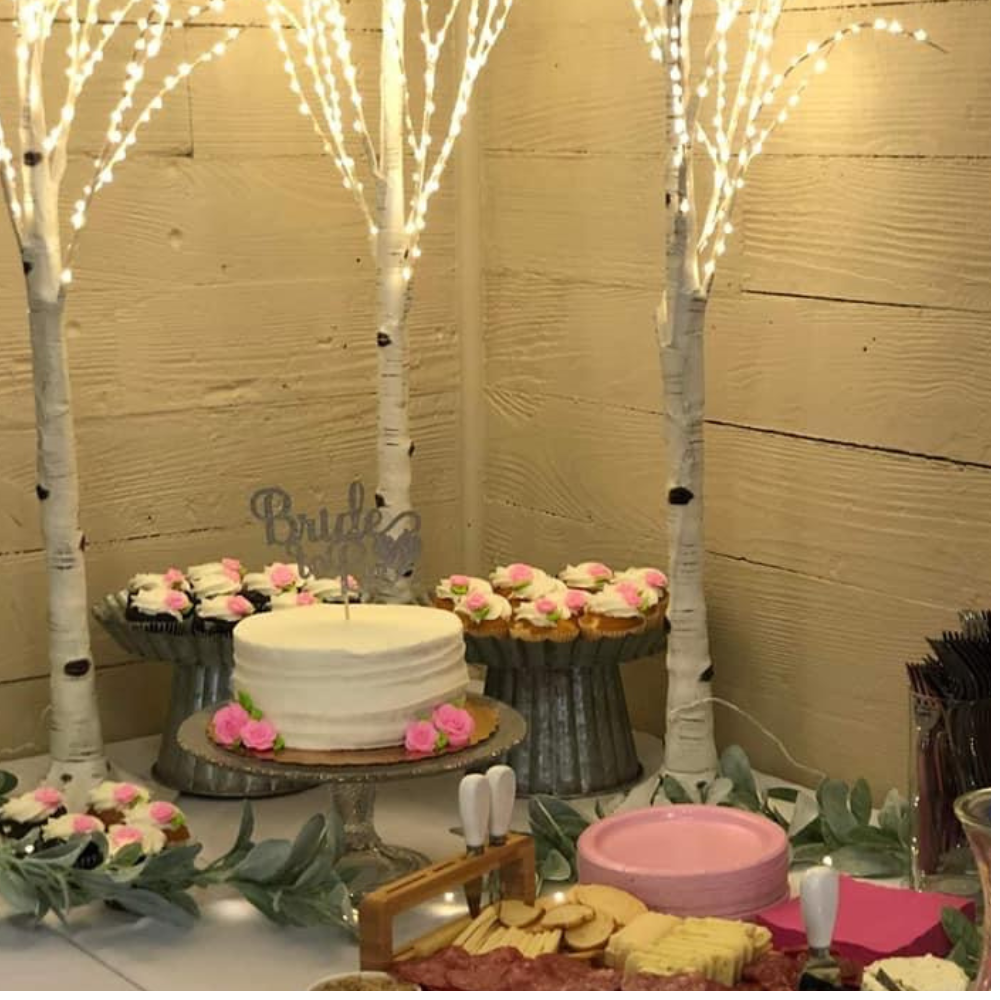 Decorative dessert table with a white cake, cupcakes, and pink flowers, set against a wooden wall with illuminated white trees.