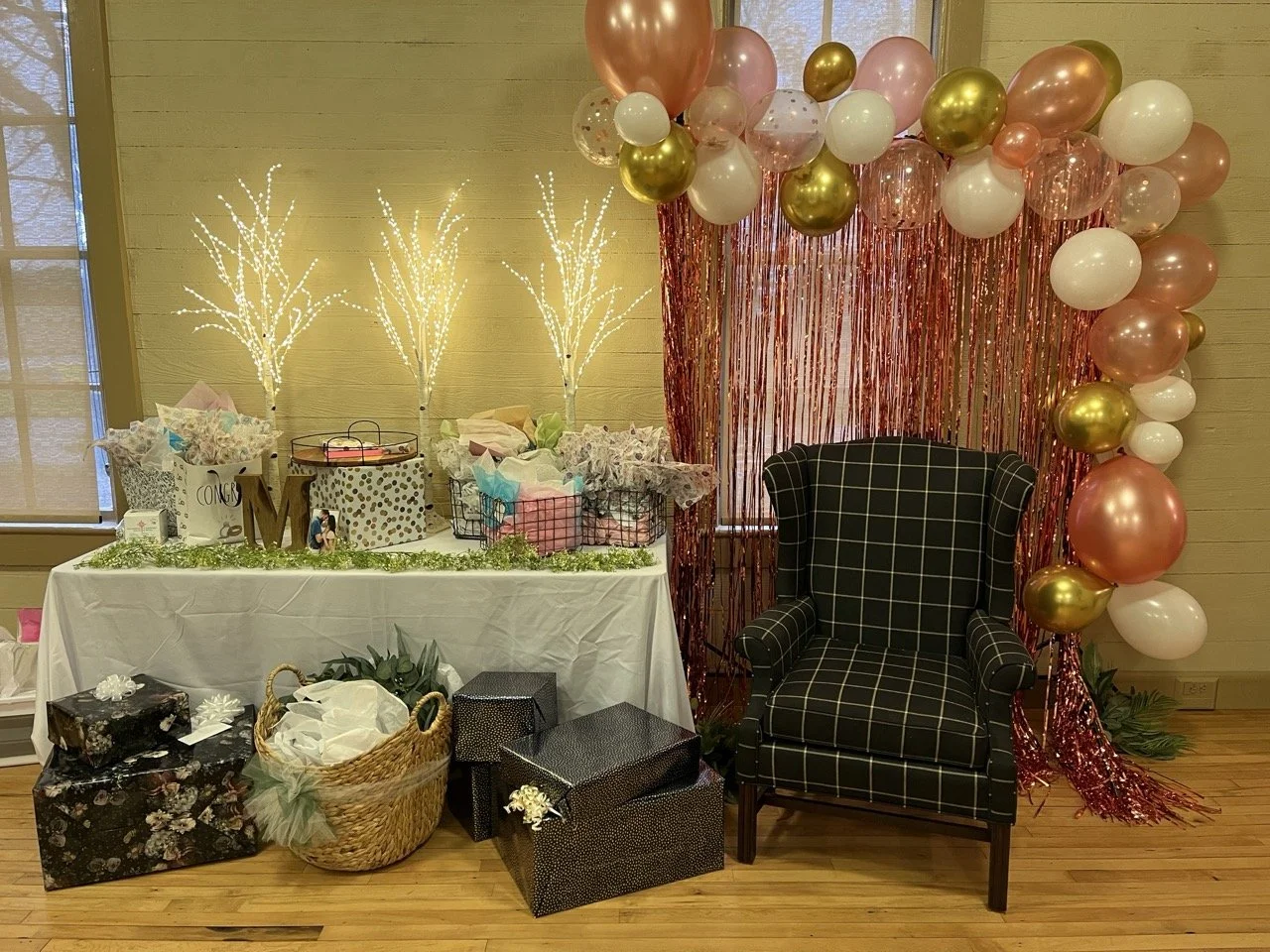 Decorations for a celebration or party with a balloon arch, gift boxes, a conga bag, and a black armchair in front of a pink metallic fringe backdrop.