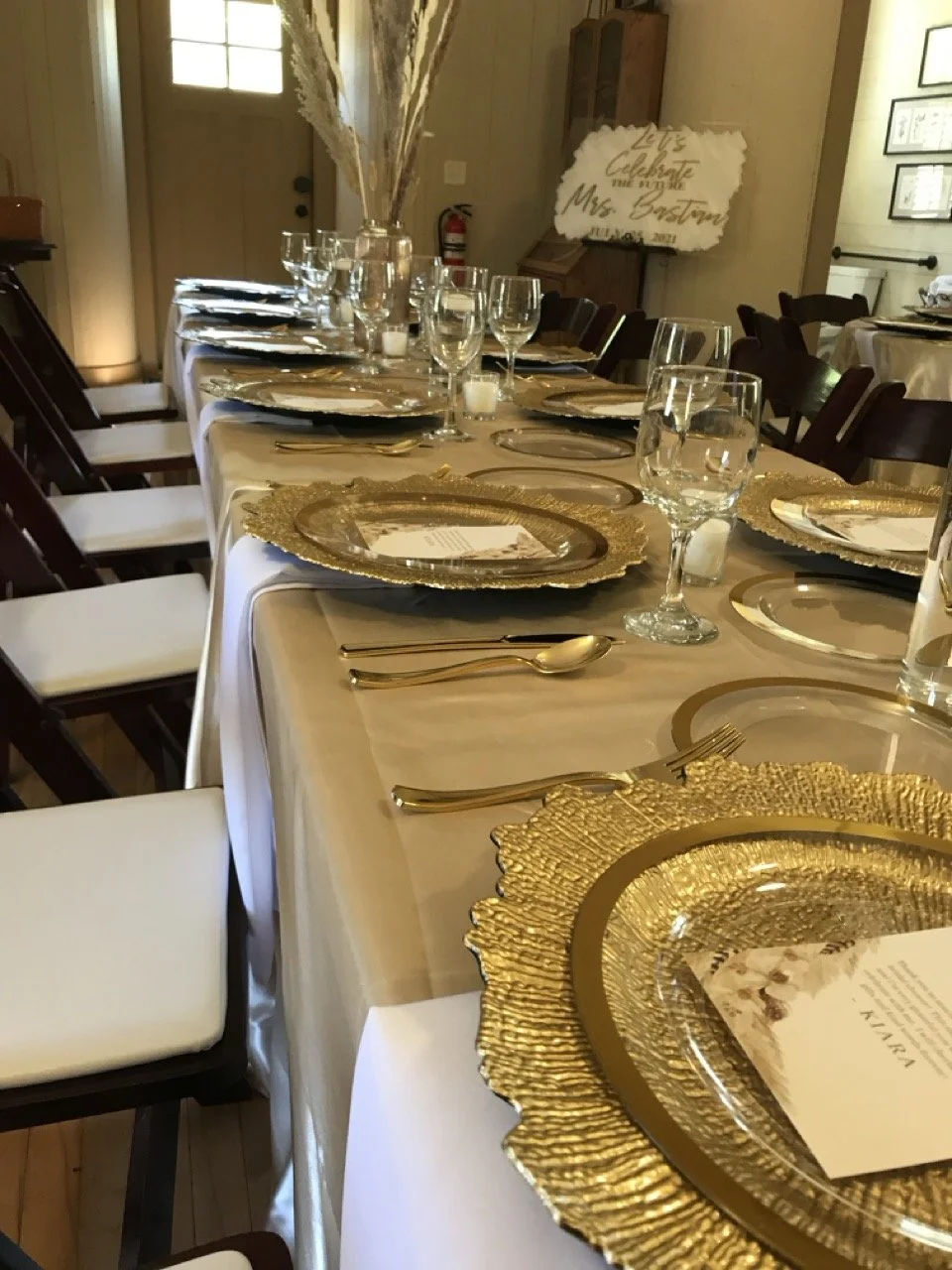 Arranged dining table with gold charger plates and gold utensils, clear wine glasses, and white napkins in a warmly lit room, decorated for a special event or celebration.