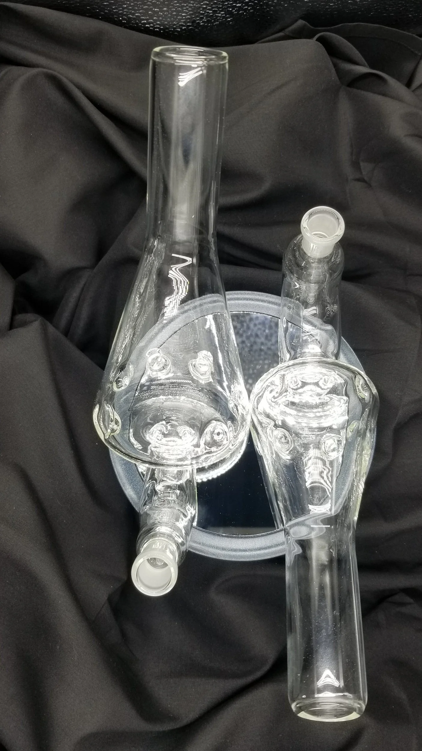 Two clear glass water pipes or bongs placed on a metallic tray, with a black cloth background.
