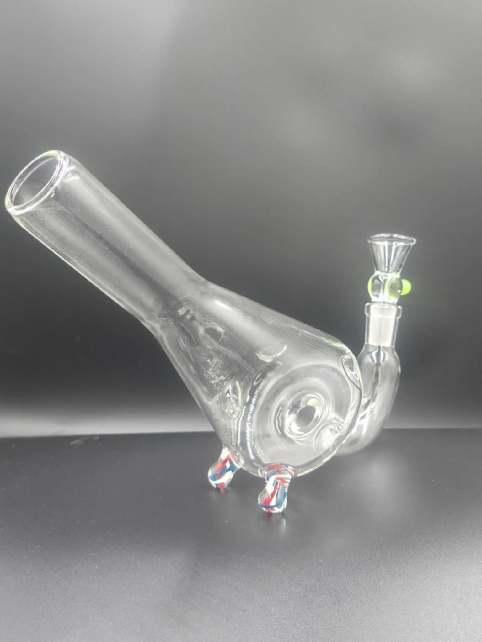 Clear glass pipe with, featuring two small feet for standing, and a green accent near the bowl, set against a dark gray background.