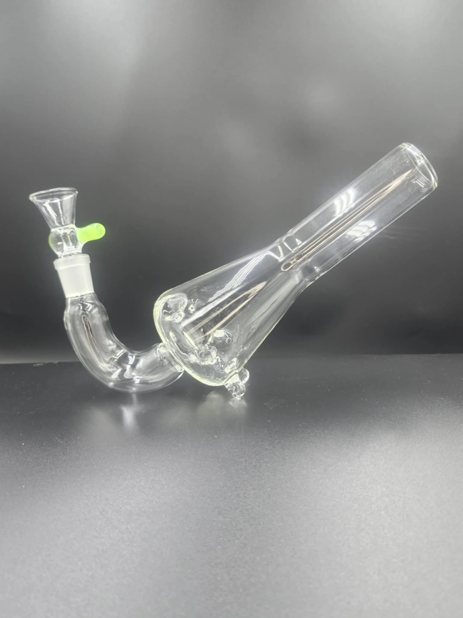 A clear glass smoking pipe with a green bowl and a curved tube design, placed on a black surface against a dark background.