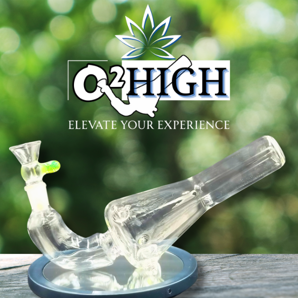 A glass weed pipe on a round black stand with a green glass bowl, placed on a wooden surface. The background is blurred green foliage. The logo says 'O2 HIGH' with a cannabis leaf and the slogan 'Elevate Your Experience' beneath.