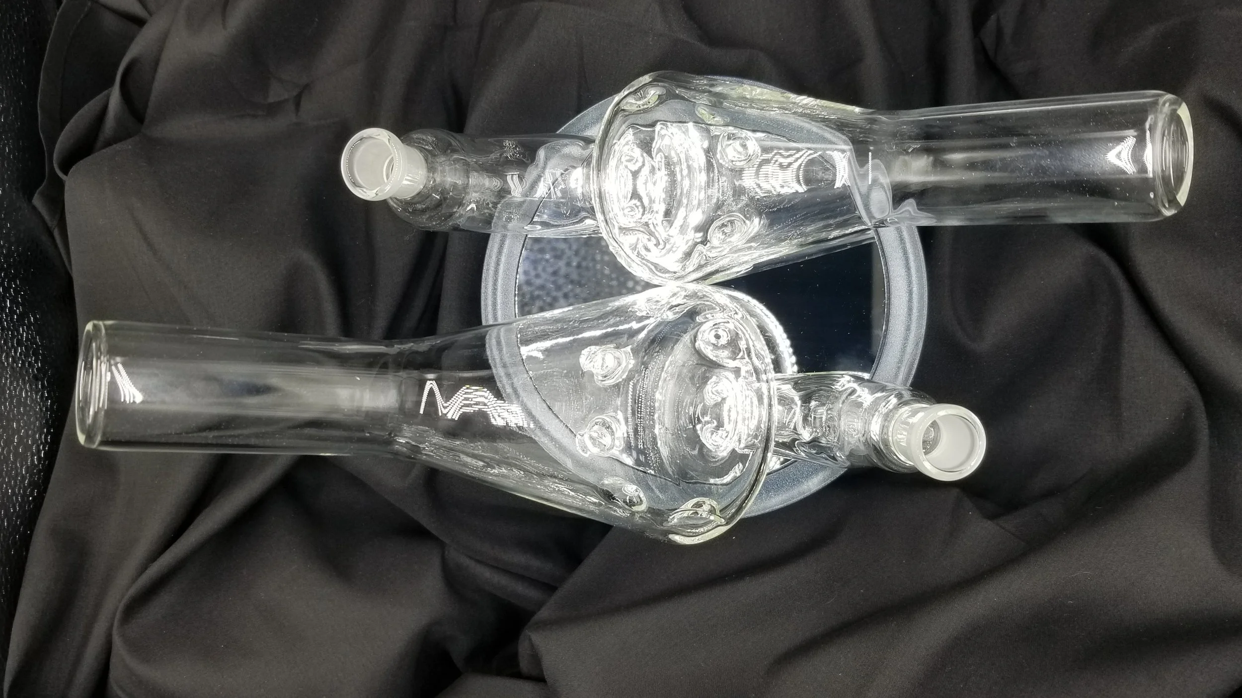 Two empty pipes with clear glass and straight tubes, resting on a reflective surface, placed on black fabric.