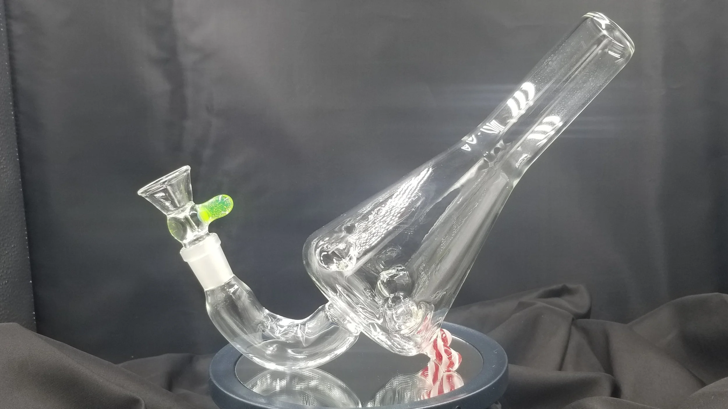 Clear glass smoking pipe with a green accent on the bowl, resting on a black surface with a black background.