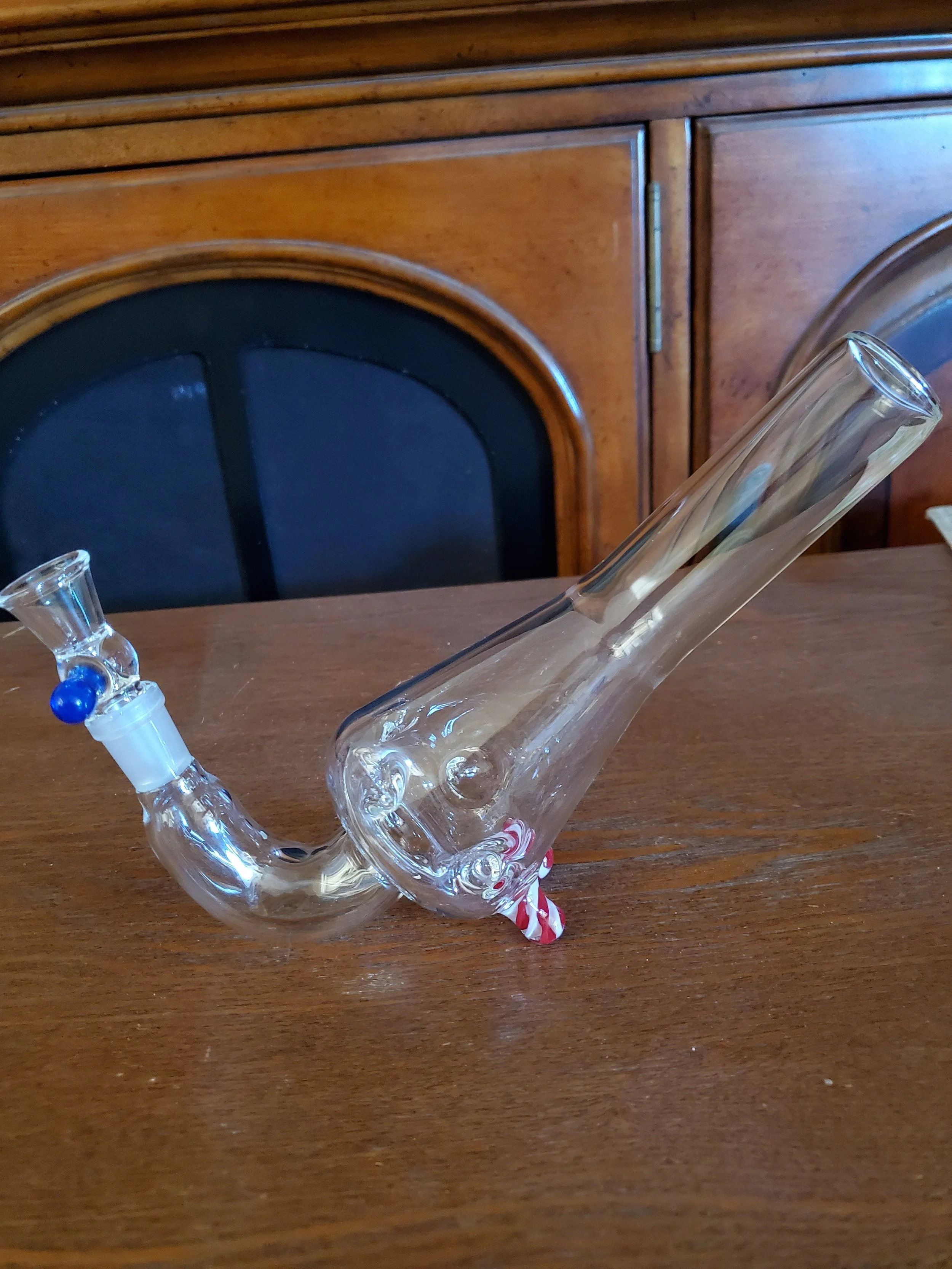 A glass pipe with a blue bowl is positioned on a wooden table against a wooden hutch background.