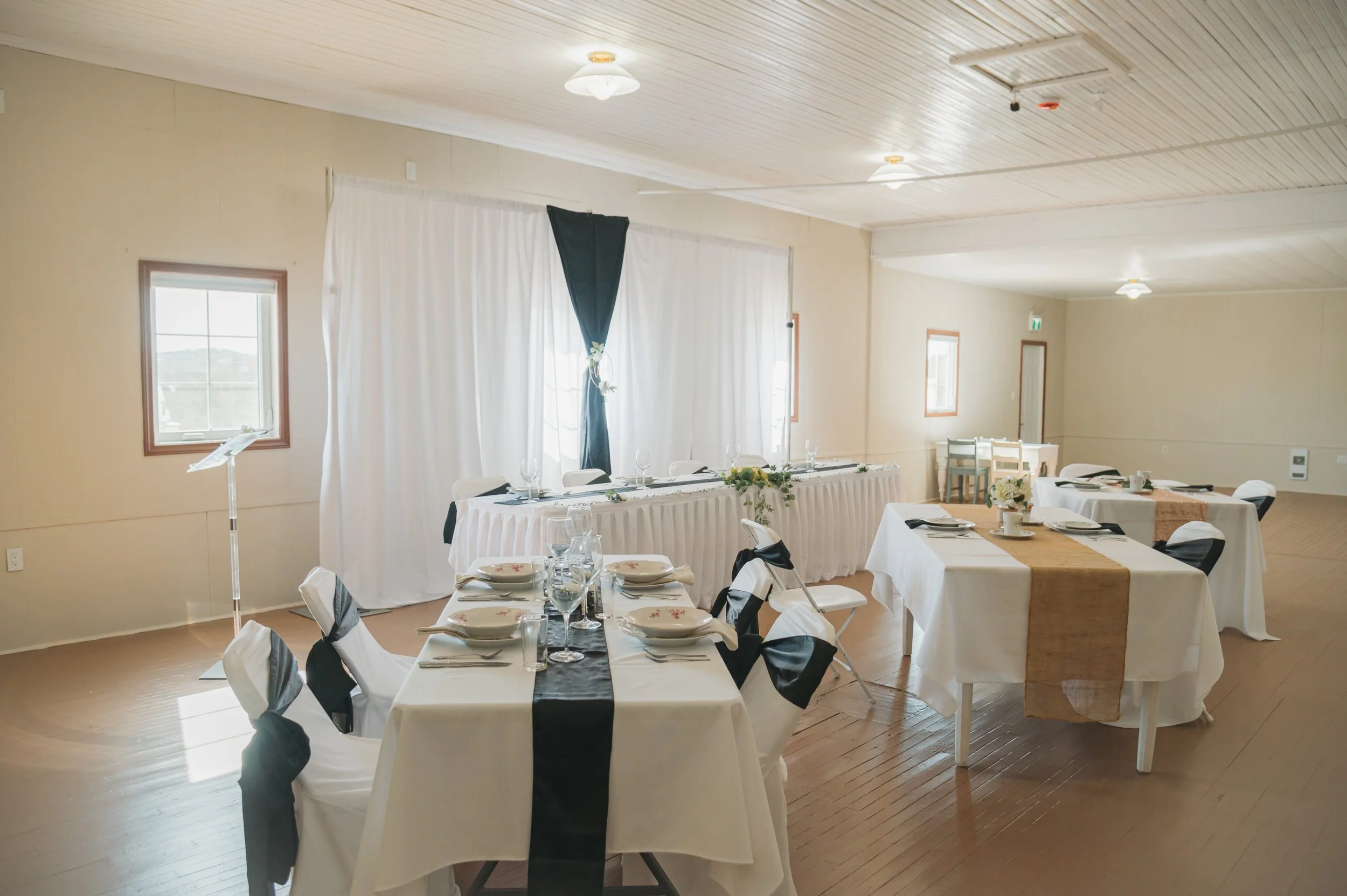 Event reception room decorated with white and black table settings, chairs with white covers and black sashes, and a head table with white tablecloth and black and white accents. There are windows and doors along the walls and a wooden ceiling.