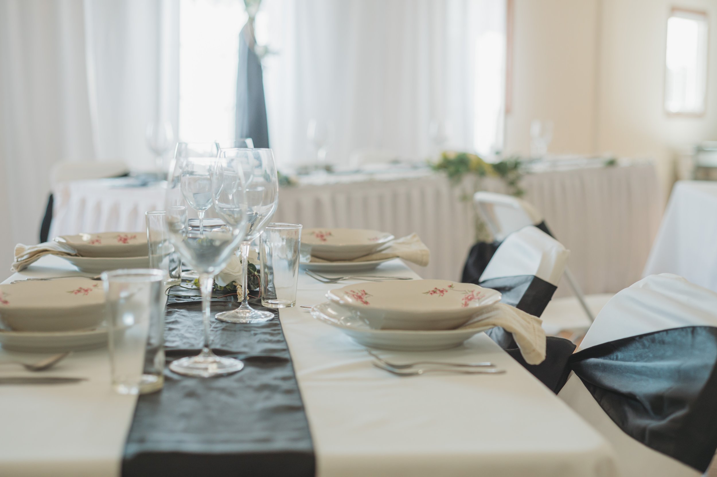 Elegantly set dining table with white plates, glasses, and black-and-white napkins at a formal event, with a long decorated table visible in the background.