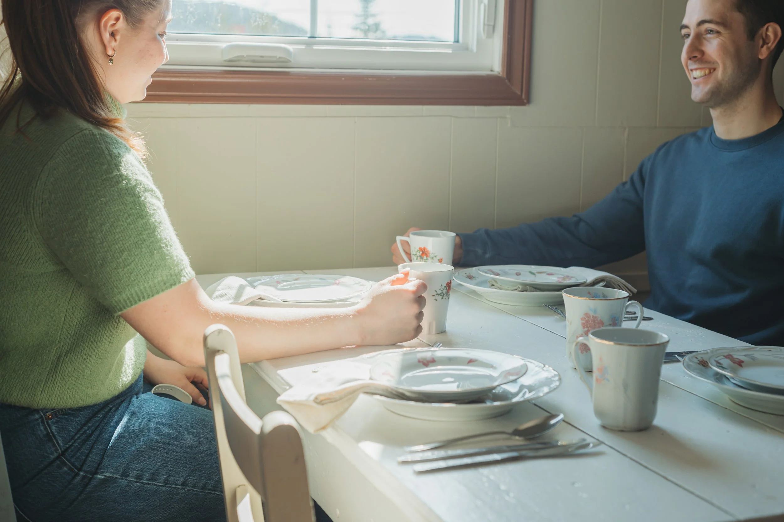 A woman and a man sitting at a dining table set with plates, cups, and silverware, enjoying a meal and smiling at each other in a sunlit room.