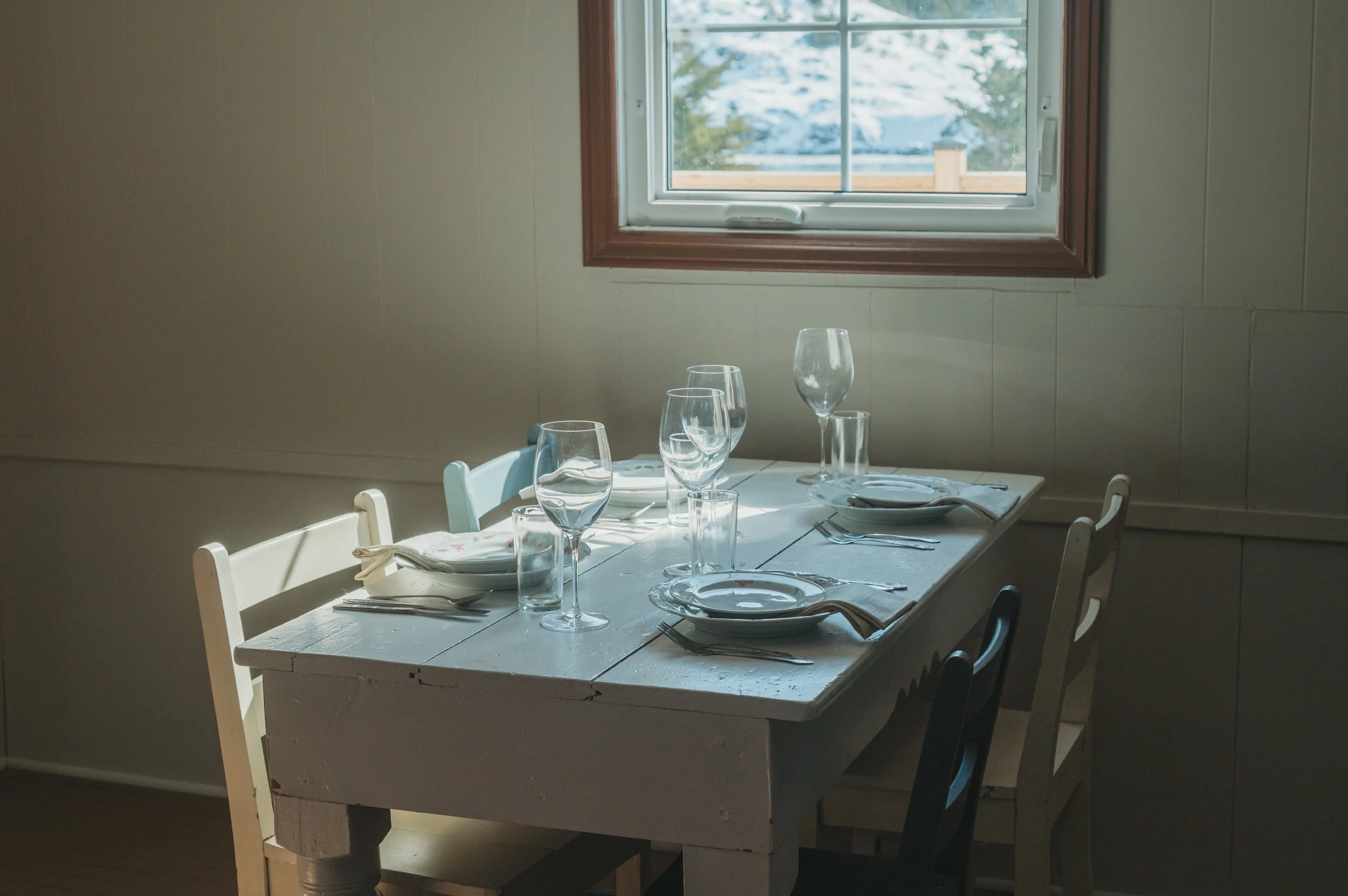 A dining table set for four with wine glasses, plates, and silverware near a window with sunlight coming through.