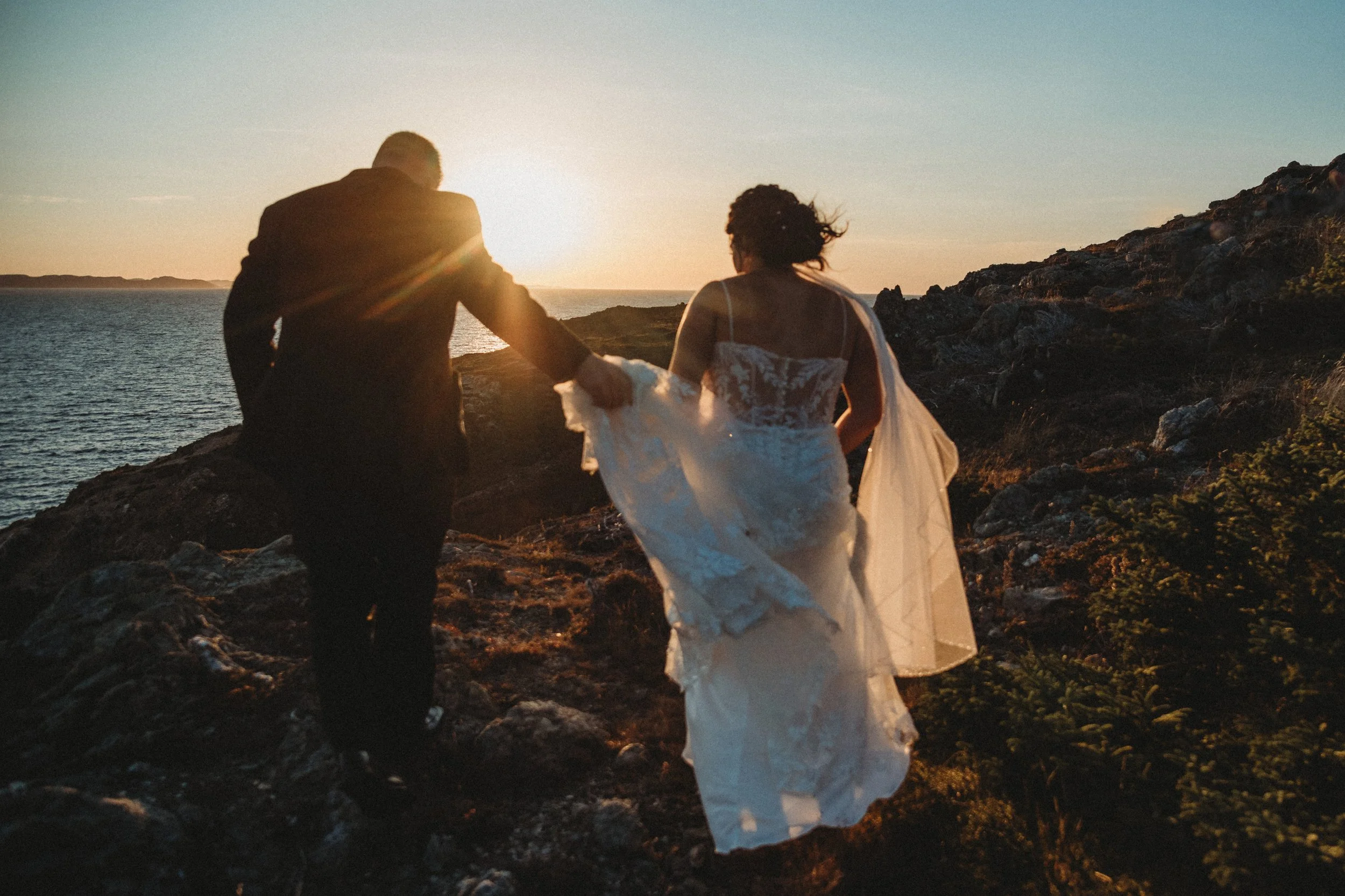 A couple in wedding attire walking along rocky terrain near the water at sunset.