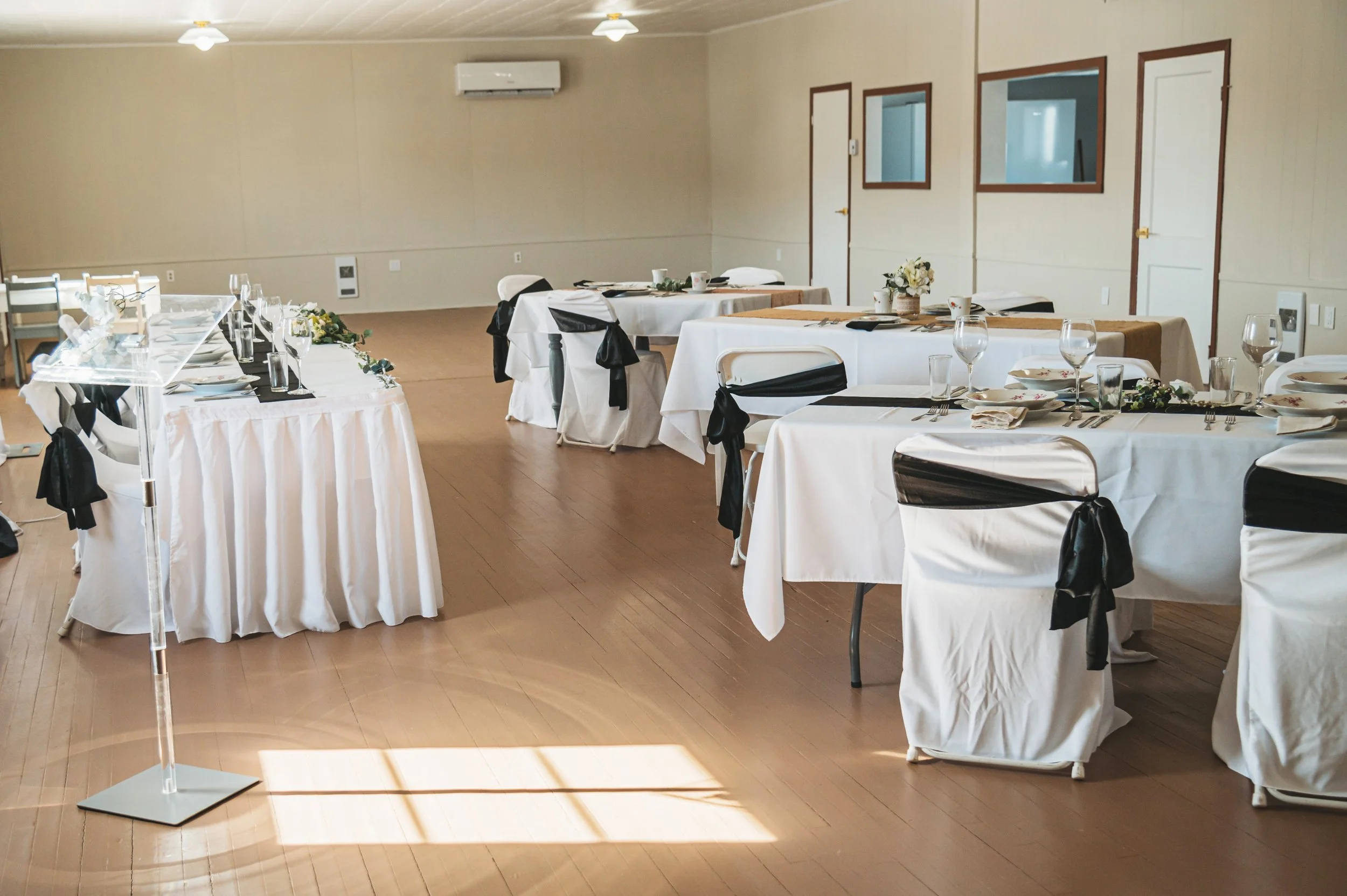Event space with tables set for a formal dinner, decorated with floral arrangements, white tablecloths, black chair sashes, and glassware.