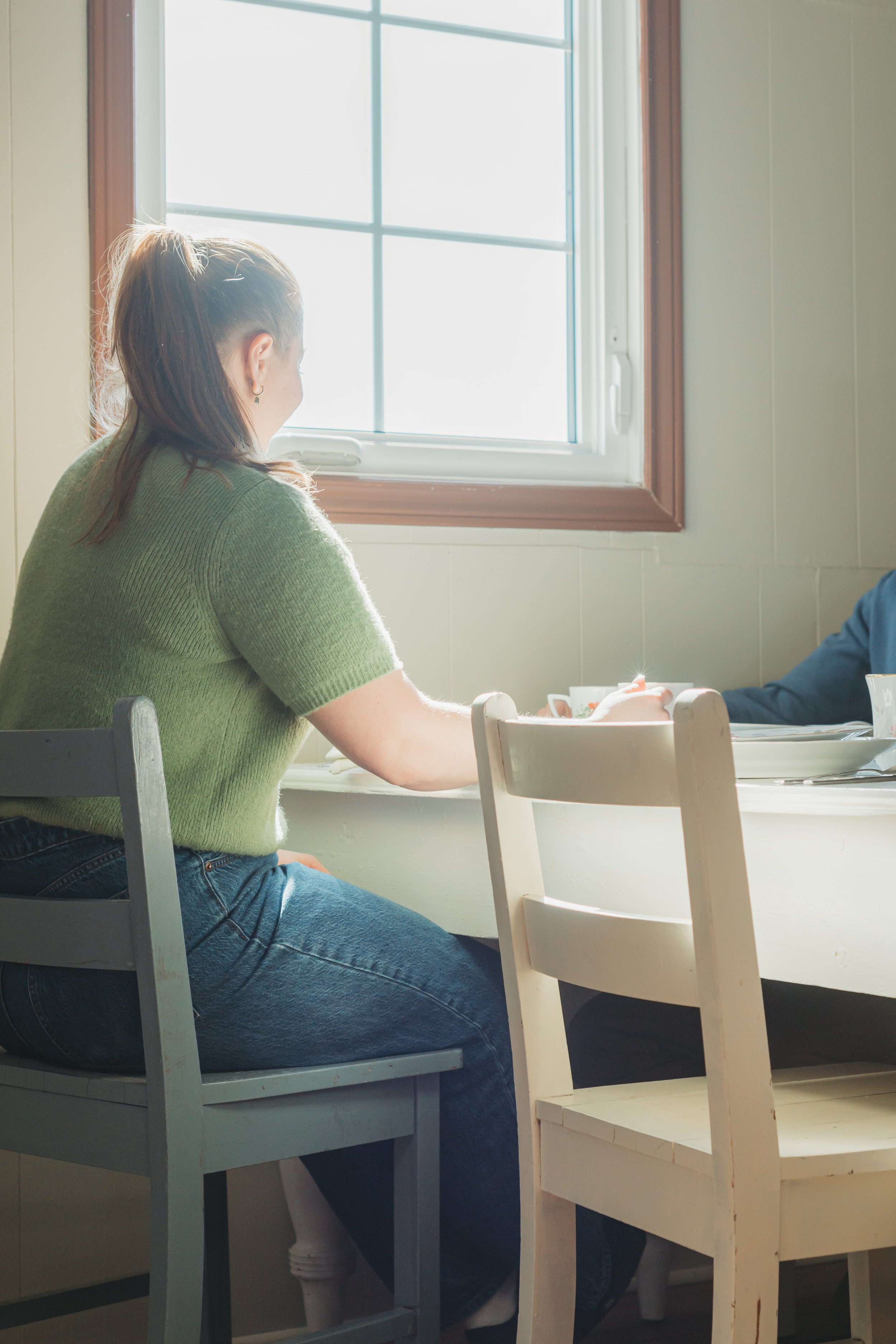 A woman with brown hair in a ponytail, wearing a green sweater and jeans, sitting by a window in a sunlit room, holding hands with someone across the table.