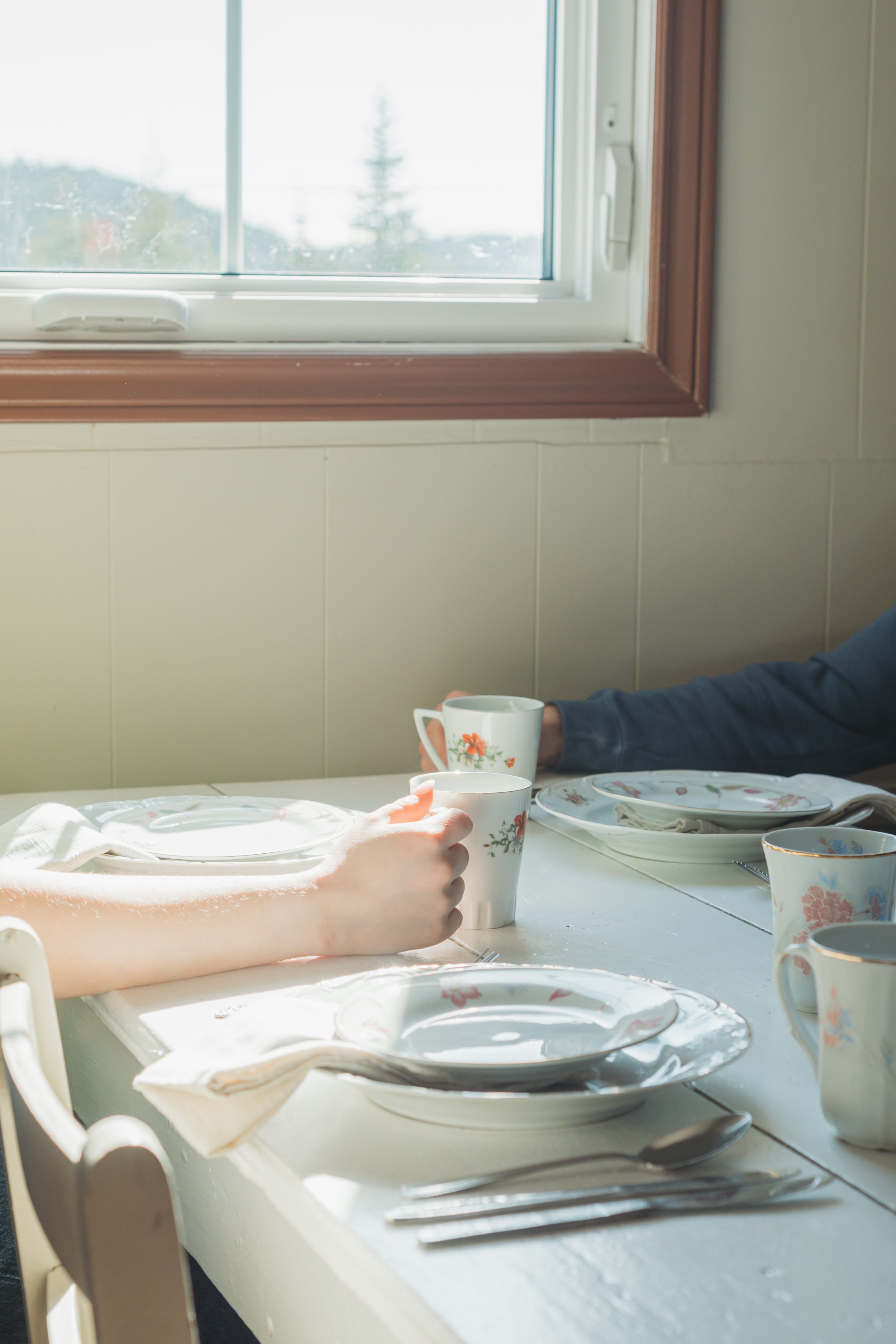 A dining table set with plates, cups, and silverware, with two people holding cups, near a window.