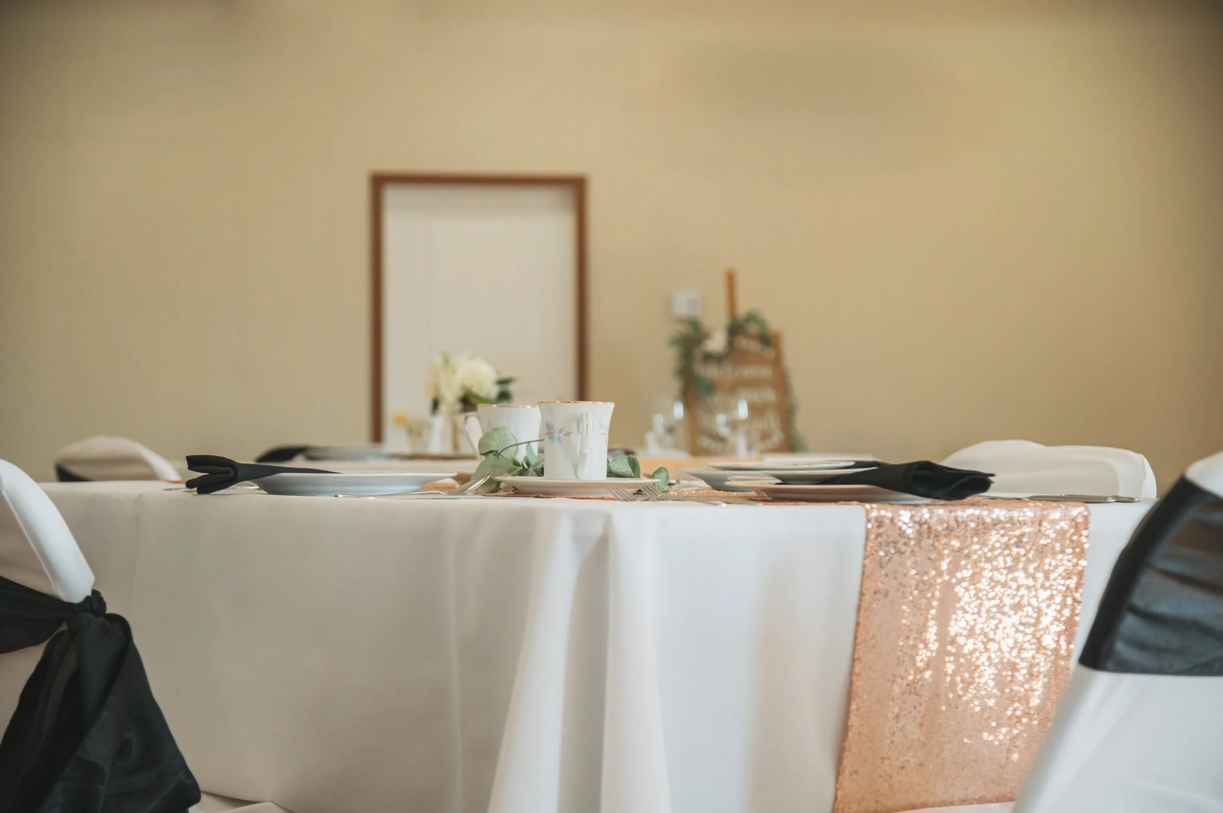 Wedding reception table decorated with white tablecloth, roses, and a rose gold sequin runner, set with plates, glasses, and cutlery, in a decorated banquet hall.