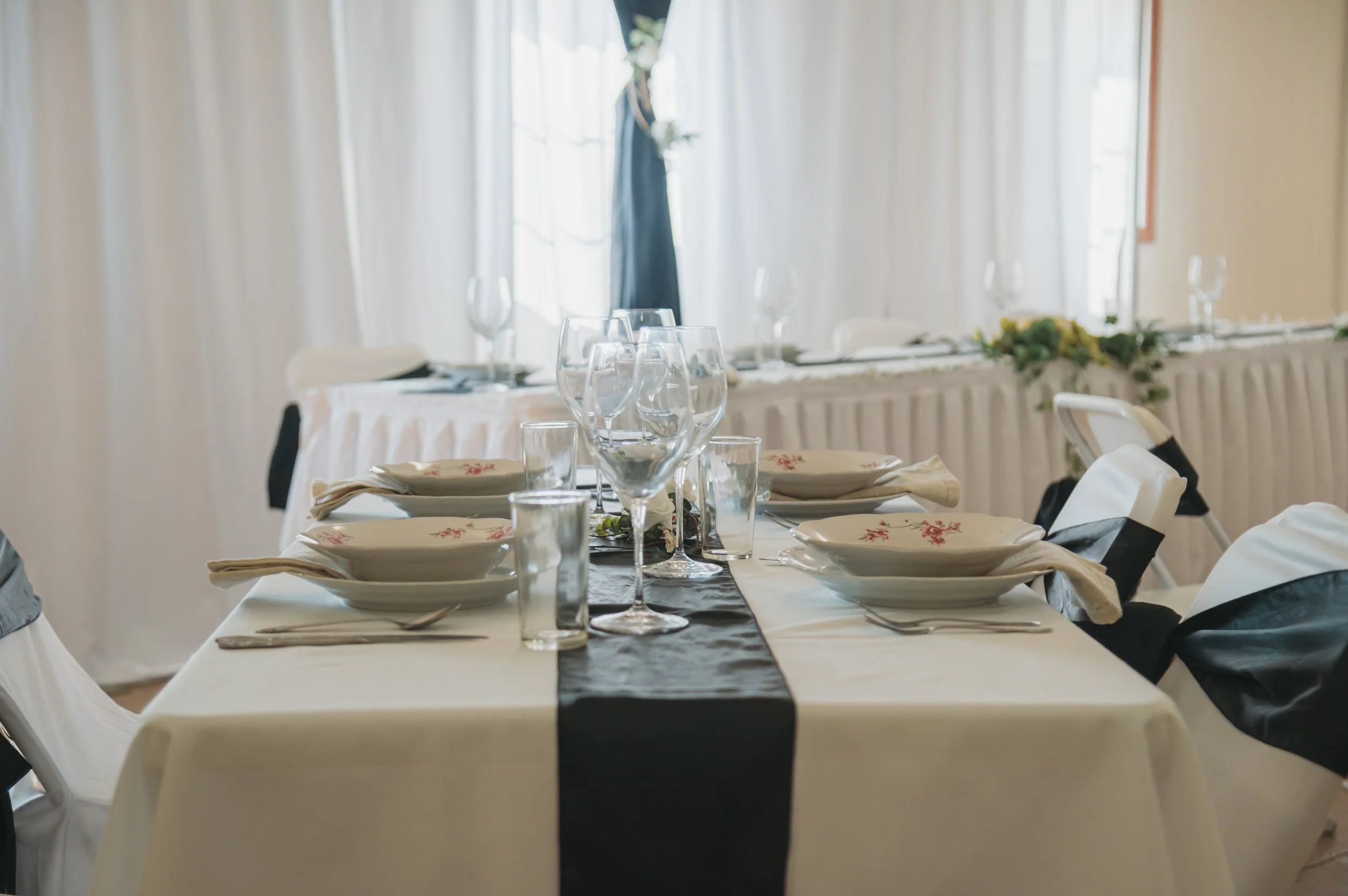 A banquet table set for a formal event with white tablecloths, black table runners, napkins, multiple wine glasses, and dinnerware in a bright room with large windows and curtains.