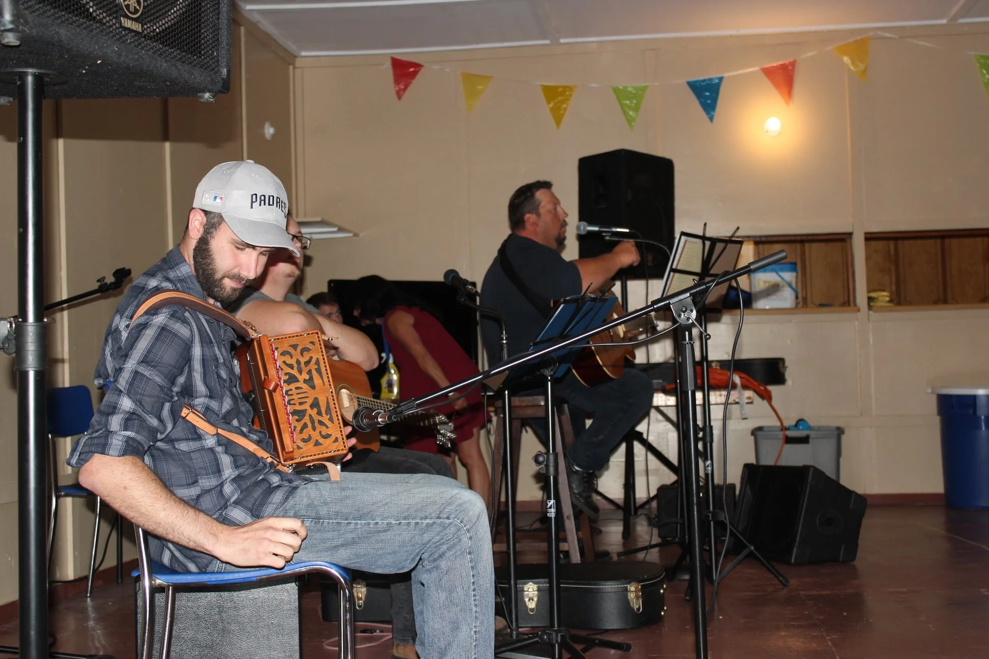 Musicians performing on stage with guitars, microphones, and music stands, colorful bunting hanging overhead, in an indoor setting.
