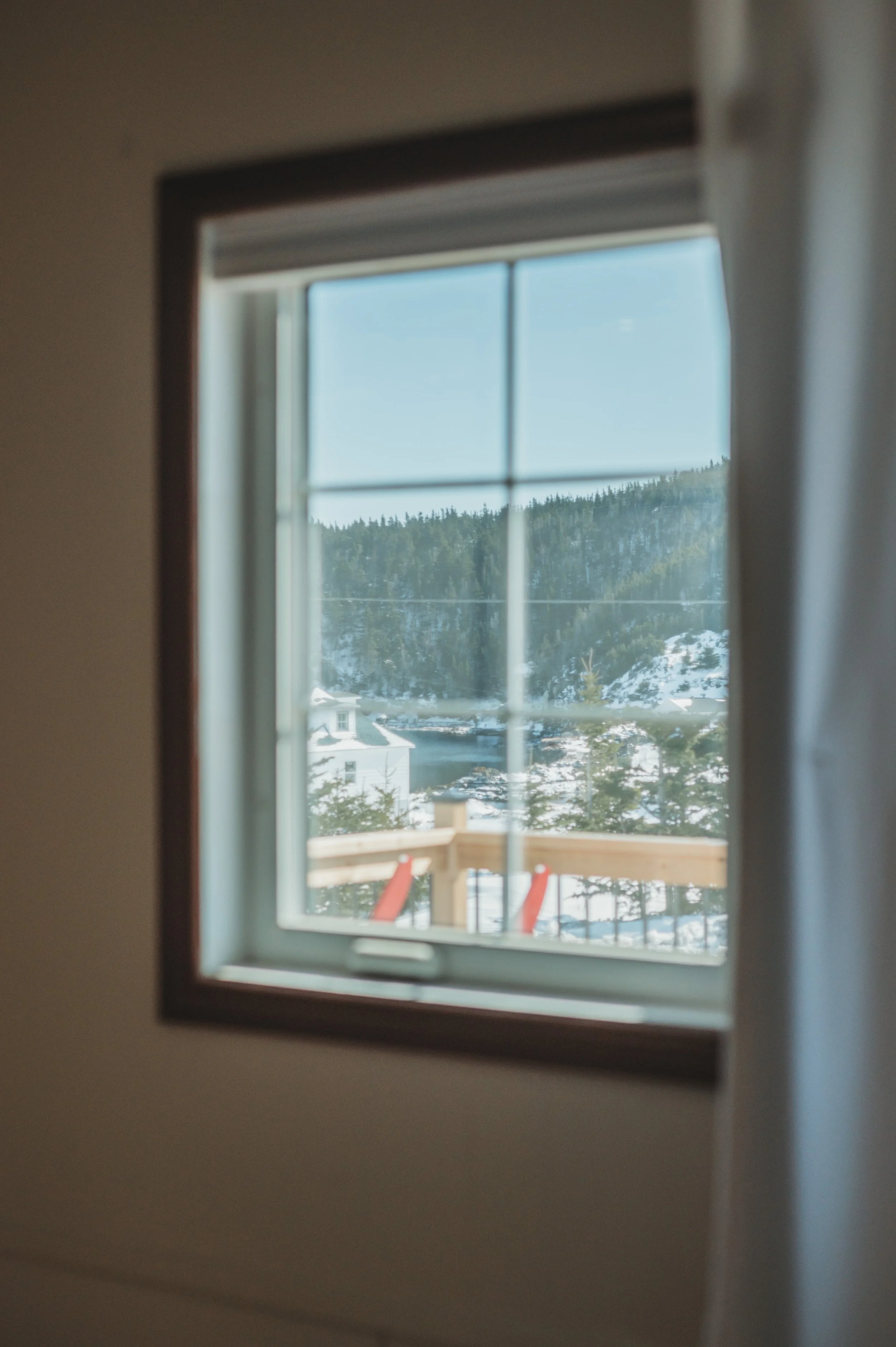 View through a window showing a snowy landscape with trees, a building, and a wooden railing outside.