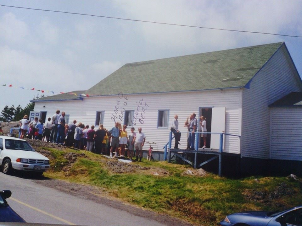 A line of people standing outside a white building with a green roof, waiting to enter. Several cars parked near the roadside, with a partly cloudy sky overhead.