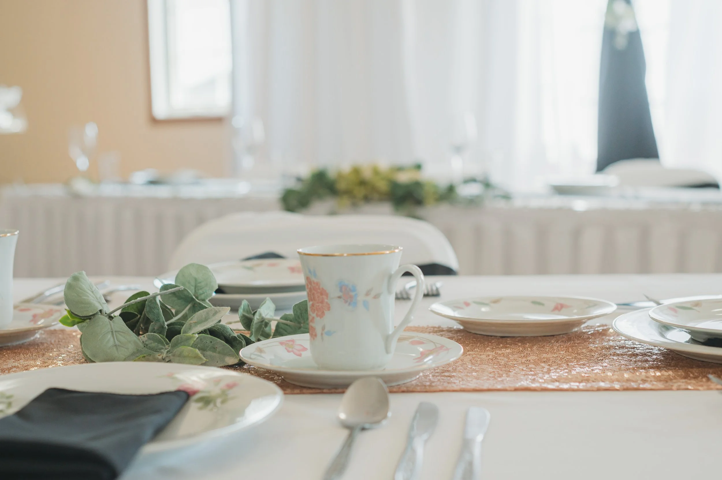 A table set for a formal meal with floral china, a gold-rimmed cup and saucer, a copper-colored table runner, and greenery centerpiece, with a softly lit, elegant dining room in the background.