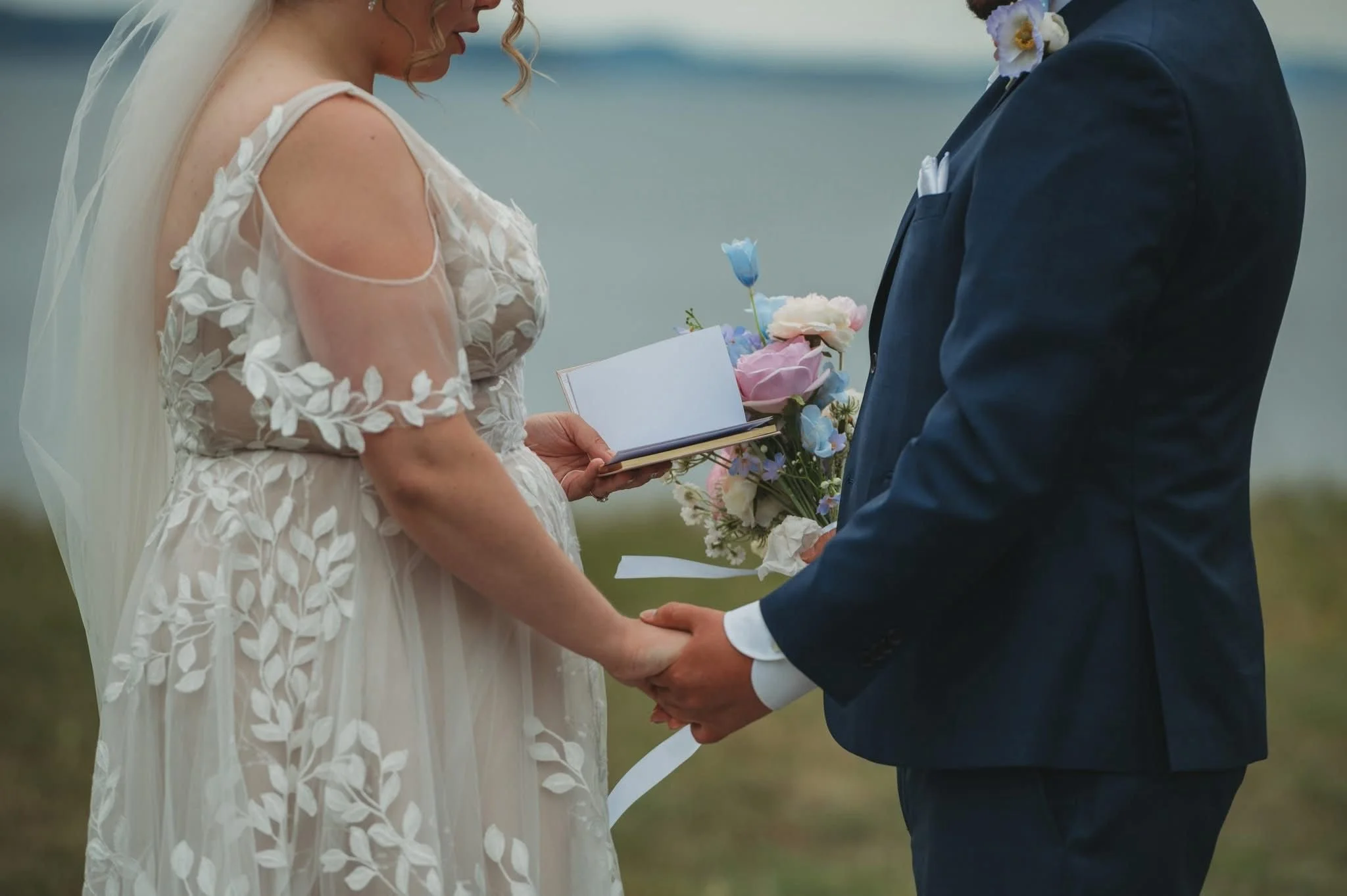 A bride and groom stand outdoors during their wedding ceremony, holding hands, with the bride reading vows from a book and the groom holding a bouquet of flowers.