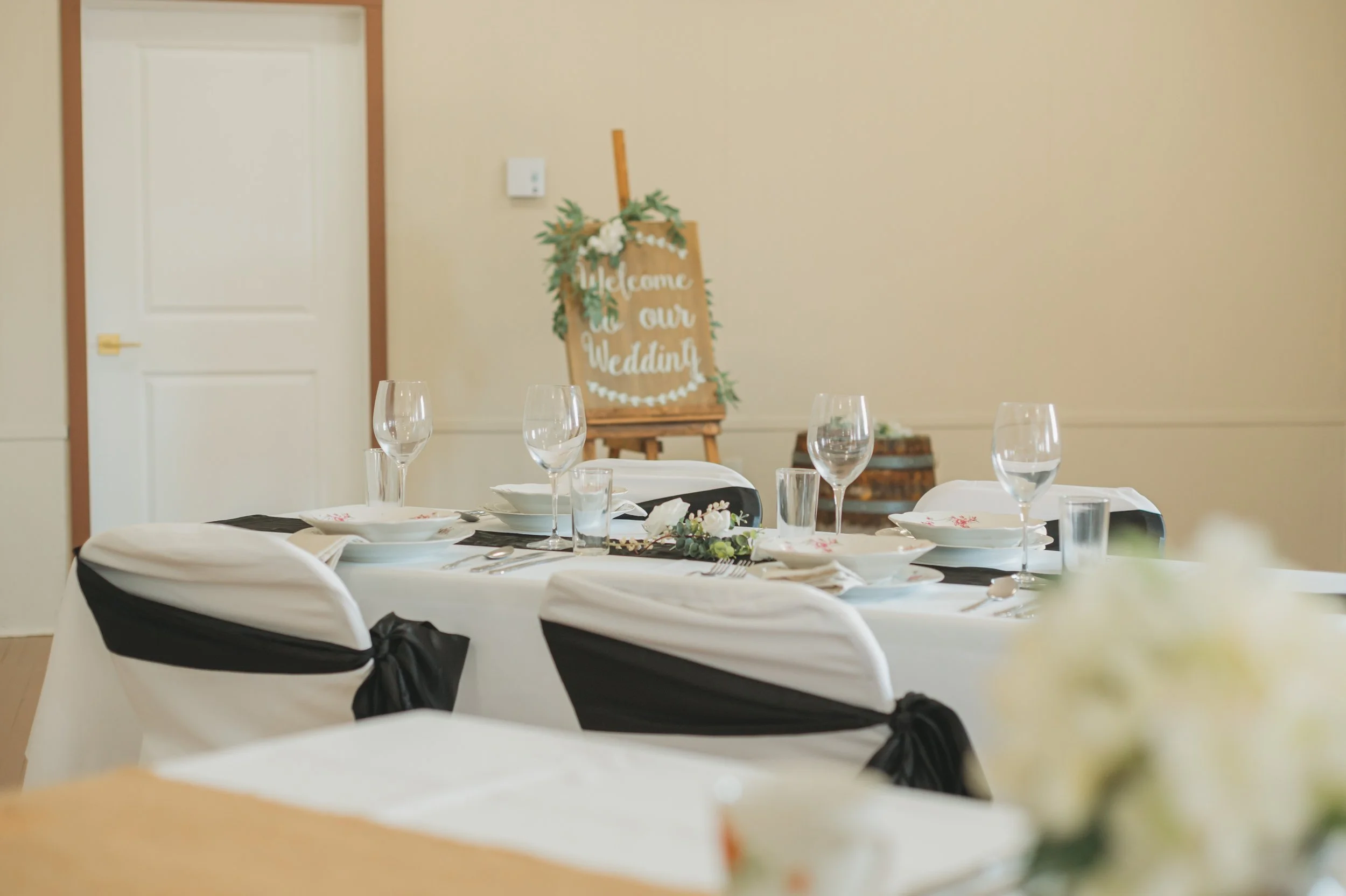 Set dining table with white tablecloth, white plates, napkins, and clear glassware, decorated for a wedding reception, with a welcome sign and greenery in the background.