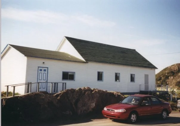 A white building with a black shingled roof, a small porch with a railing, and a blue front door, sitting on a rocky terrain with a red car parked in front.
