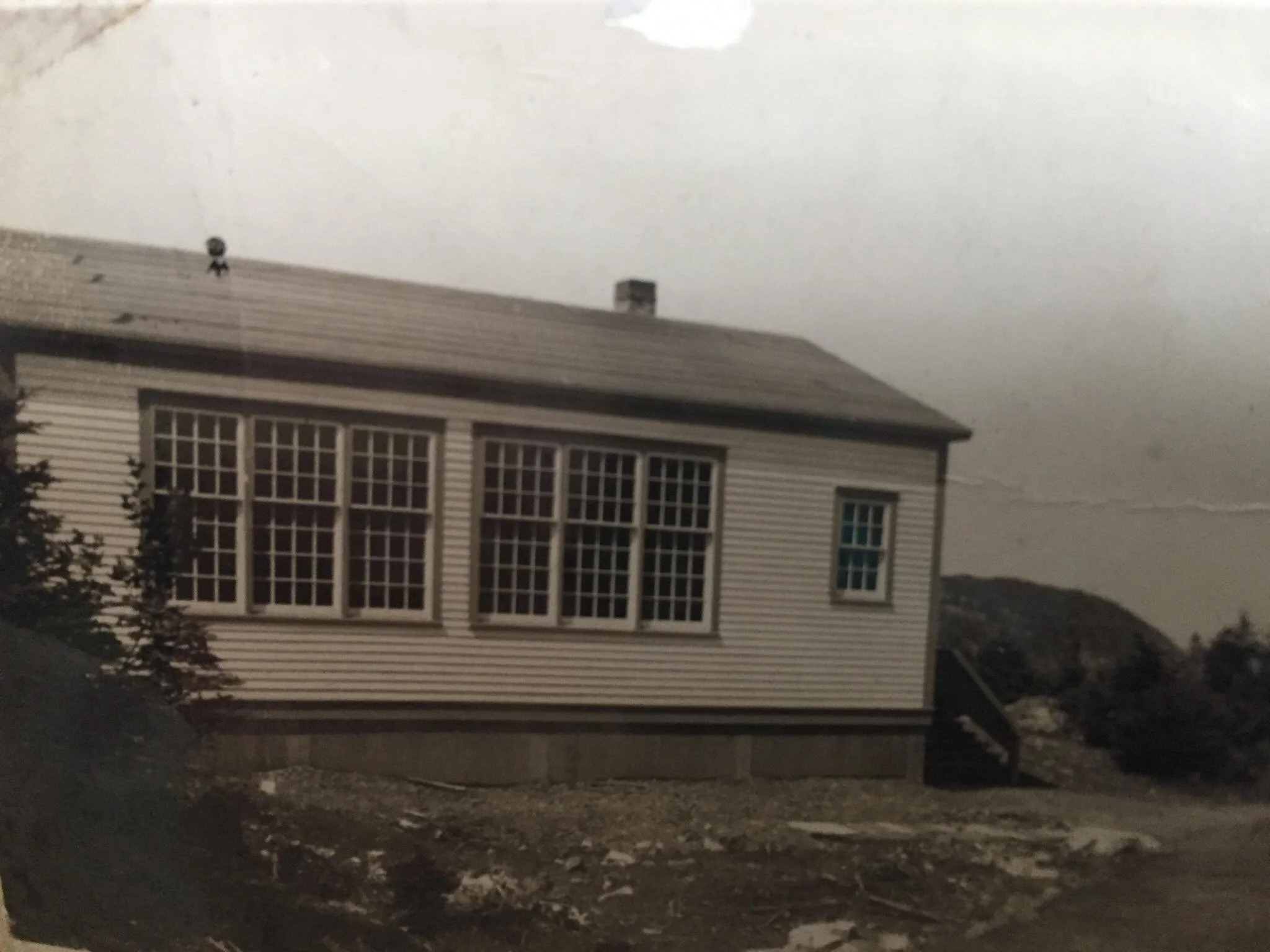 A building with a gabled roof, horizontal siding, multiple large windows, and stairs leading to the entrance, situated on uneven terrain with vegetation around.