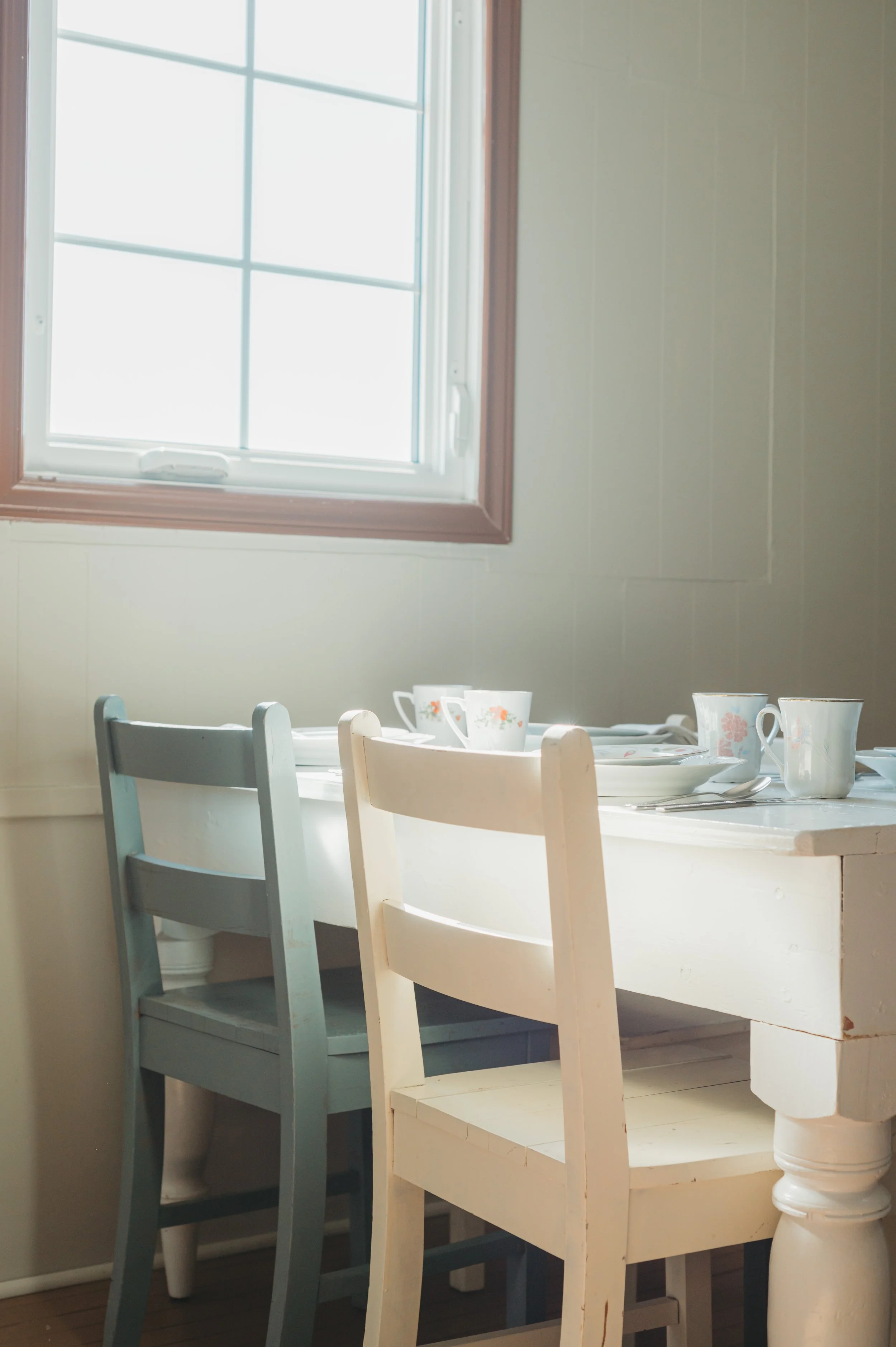 A cozy dining nook with a white wooden table, two mismatched chairs in pastel colors, and a window letting in natural light.