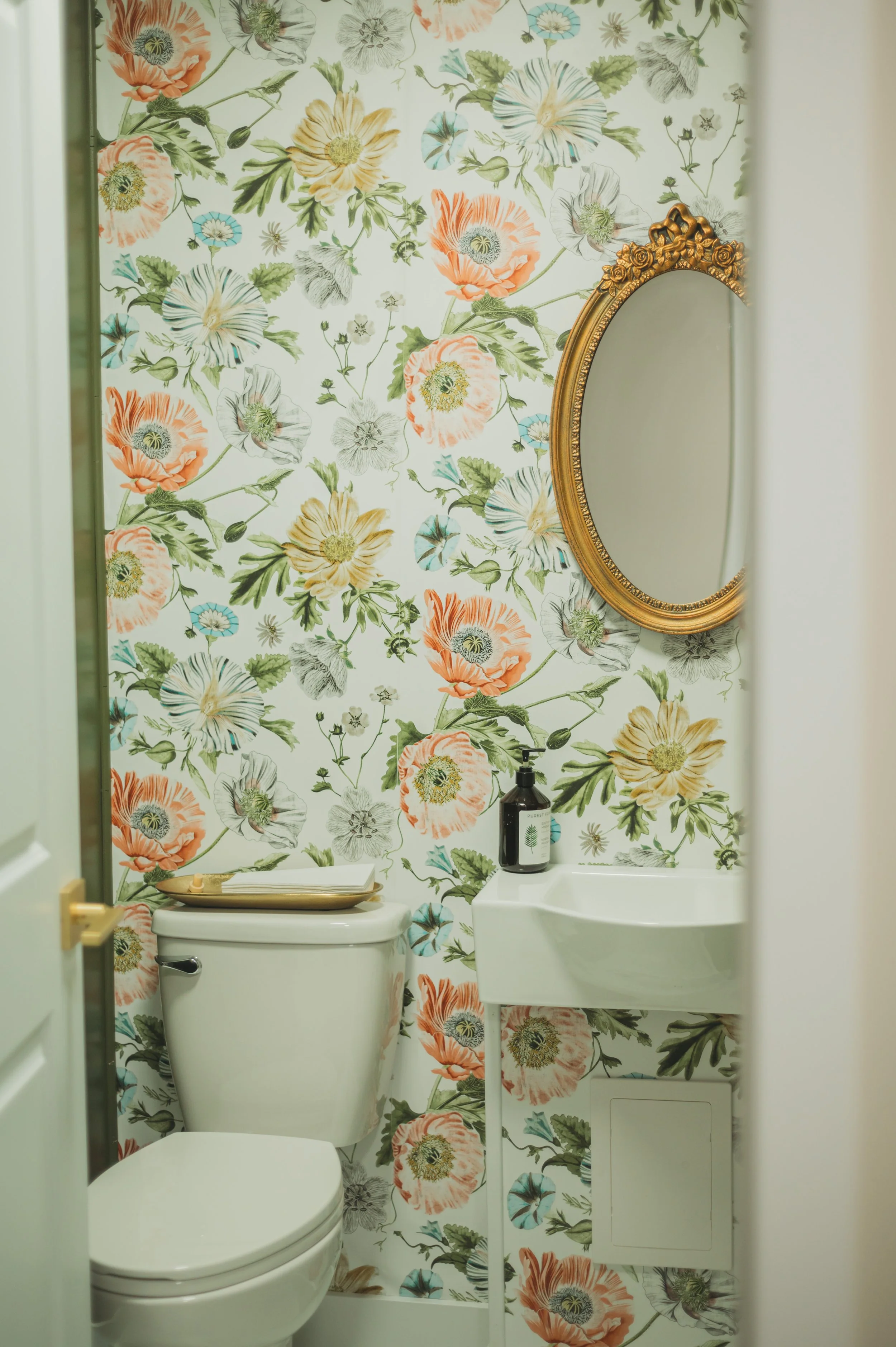 Small bathroom with floral wallpaper, a white toilet, a tiny white sink, a gold-framed mirror, and a black soap dispenser on the sink.