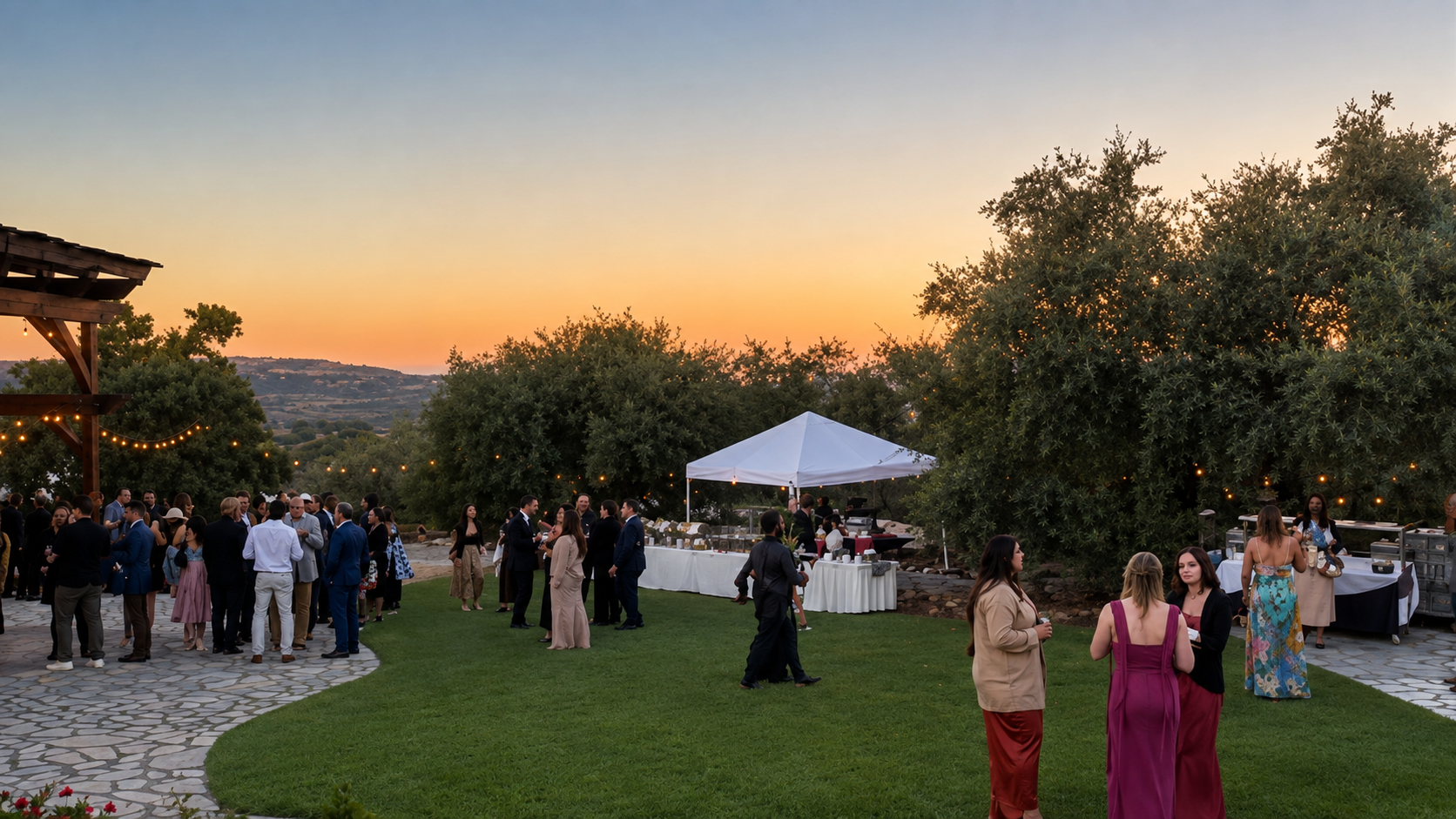 People at a social gathering outdoors during sunset, with trees, a white canopy, and string lights in the background.