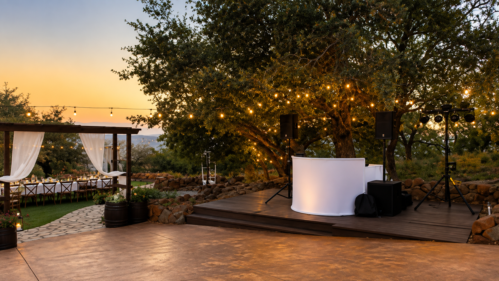 An outdoor event space at sunset with a wooden stage, DJ equipment, and string lights hanging from trees. There is a white curved table on the stage and a dining area with tables and chairs decorated with candles and flowers under a wooden pergola with white curtains.