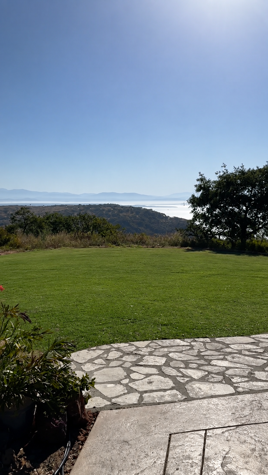 A scenic outdoor view showing a stone patio, a well-maintained green lawn, two trees on the right, and distant hills and water under a clear blue sky with the sun shining.
