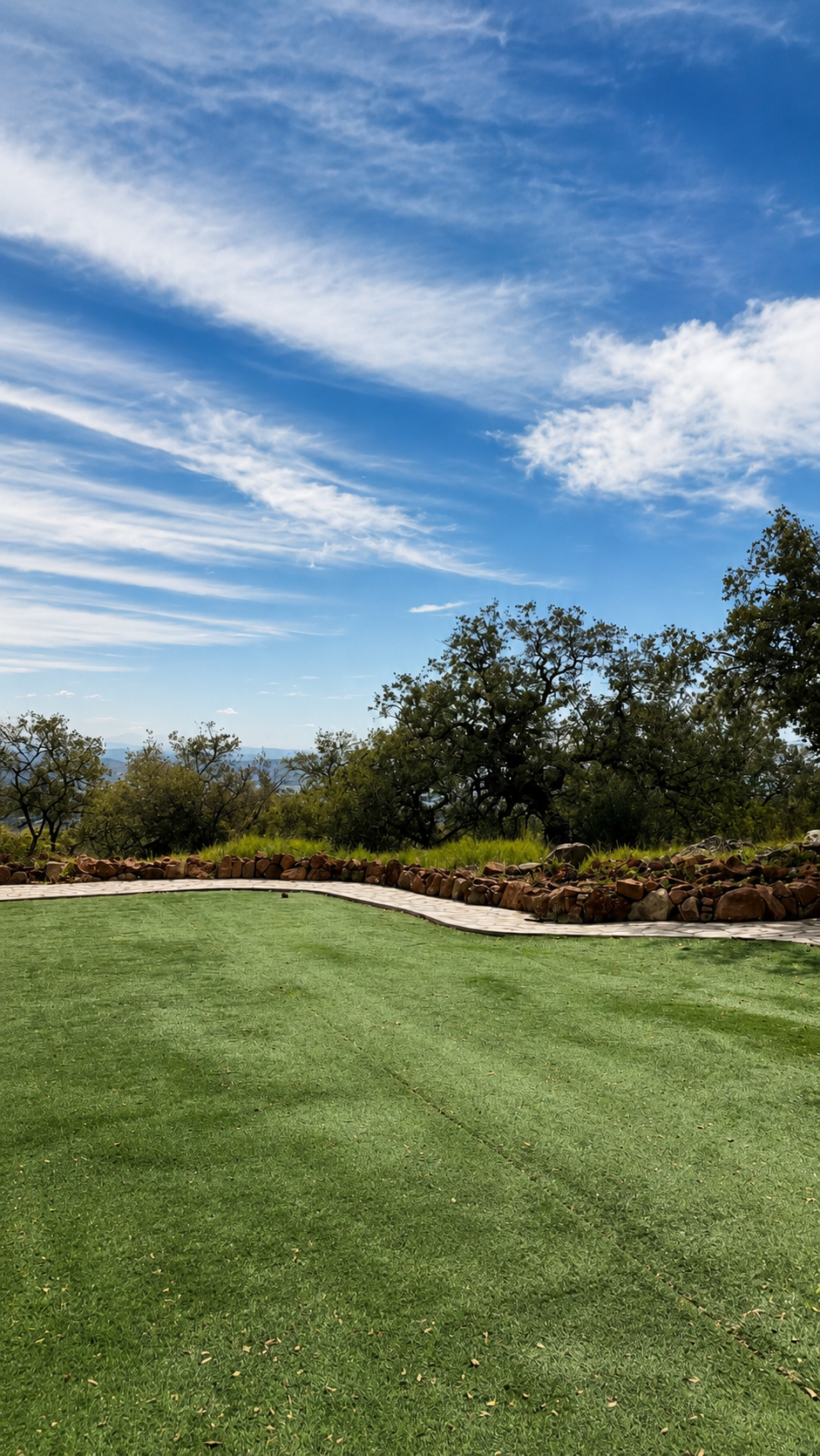 A landscaped outdoor area with a well-maintained green lawn, a stone border, trees, and a bright blue sky with clouds.