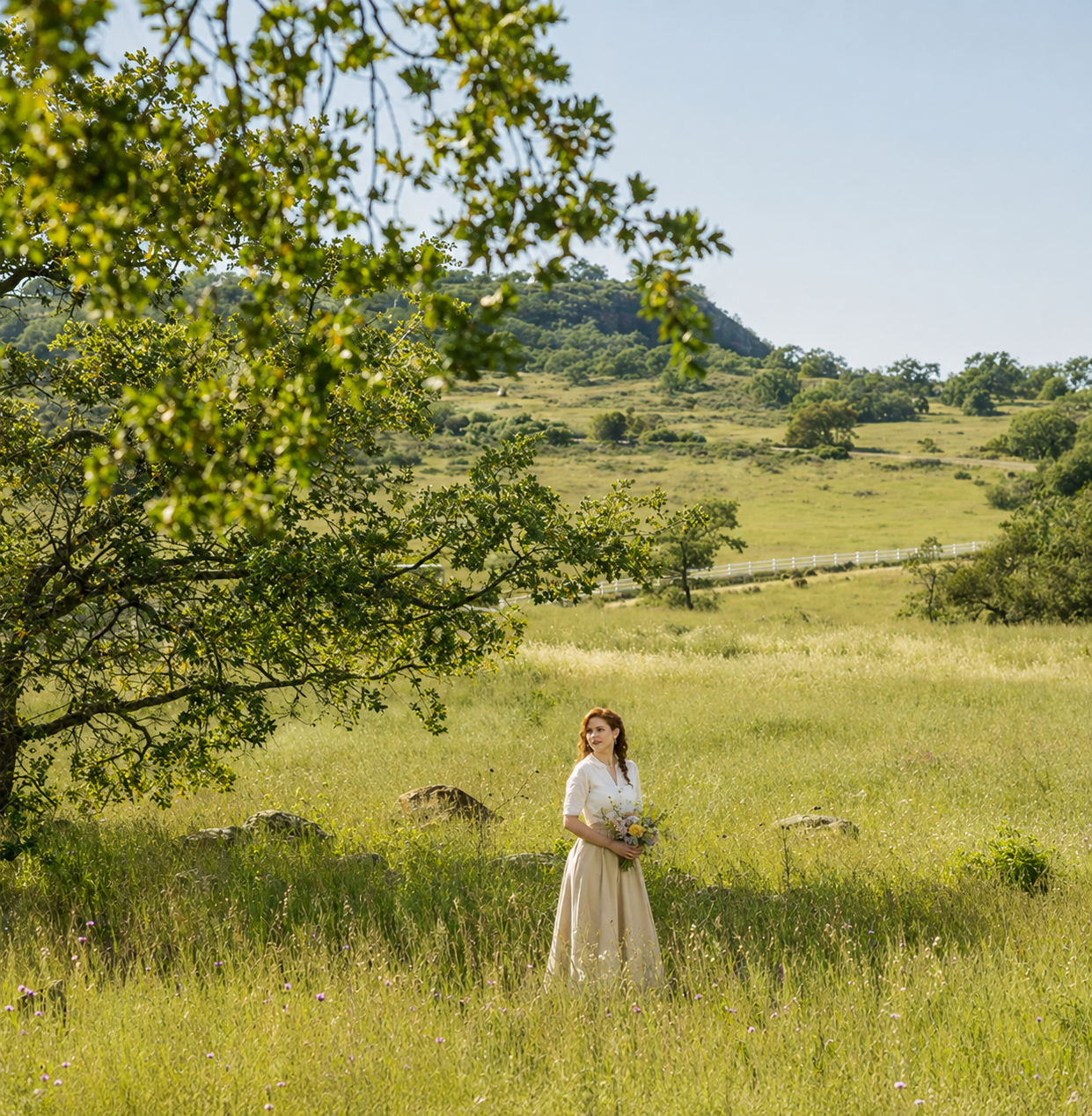 A woman in vintage attire holding a bouquet stands in a green field with trees and a mountain in the background on a bright day.