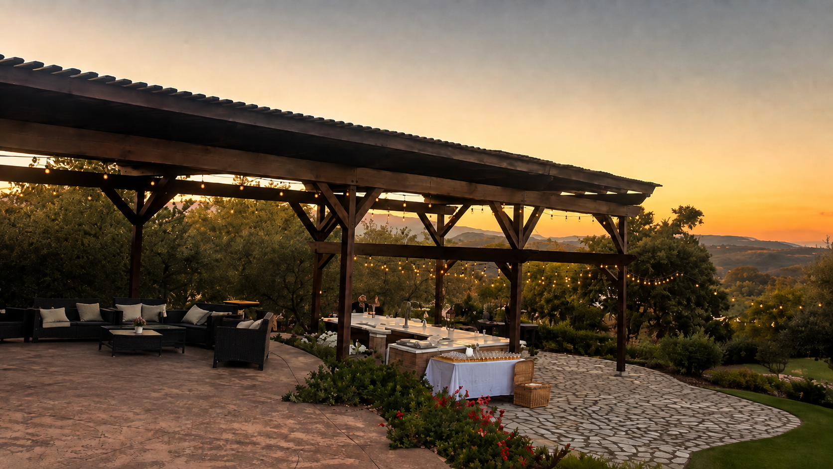 Outdoor patio area with black wicker furniture and white cushions, decorated with string lights, overlooking a scenic landscape at sunset with trees and hills.